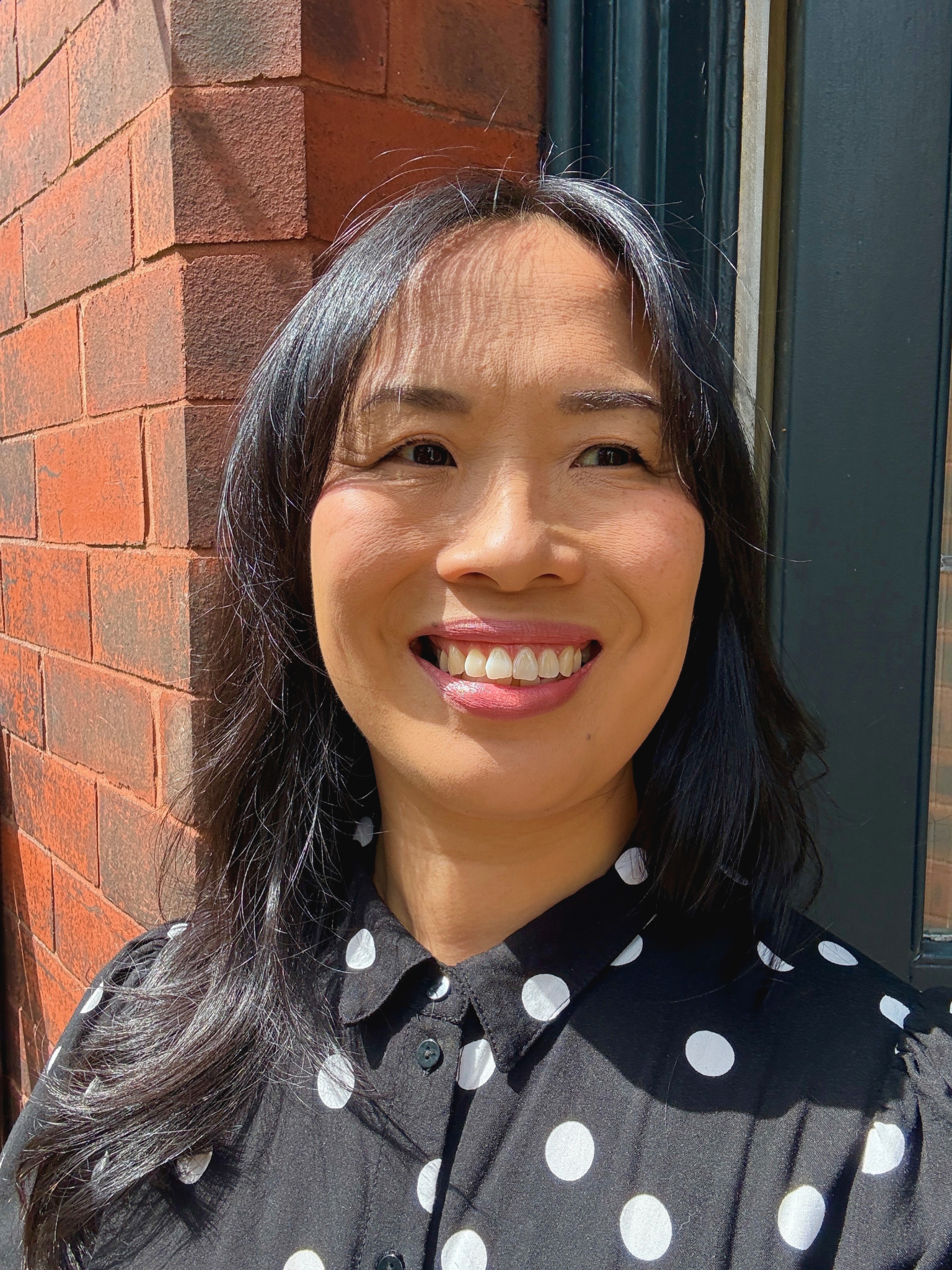Woman with long black wavy hair smiling and looking to the side. She is standing with red brick wall in the background.