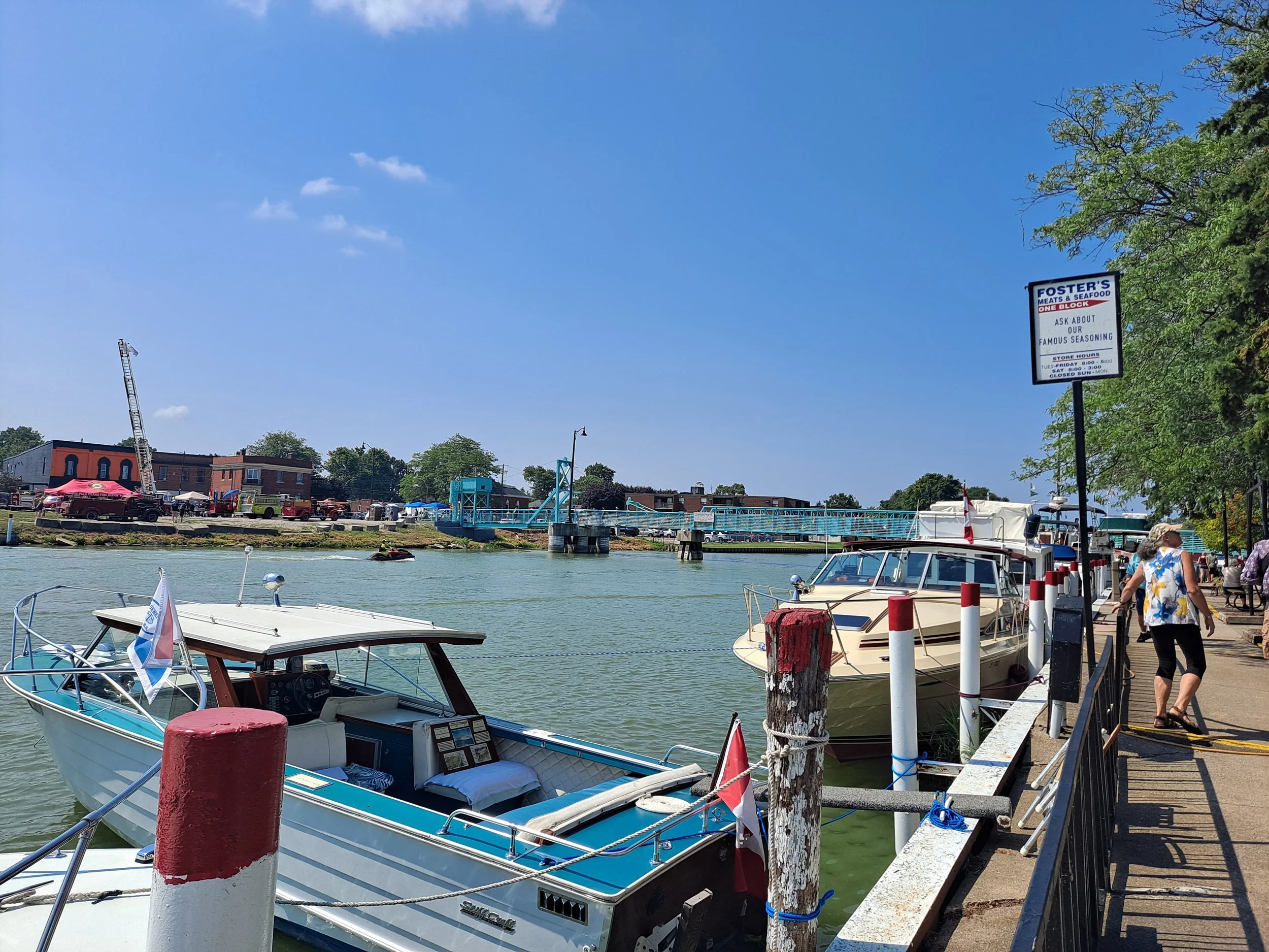 View of the Sydenham River with boats lining the docks with the walking bridge in Wallaceburg in the background which leads to the antique fire truck display in the background