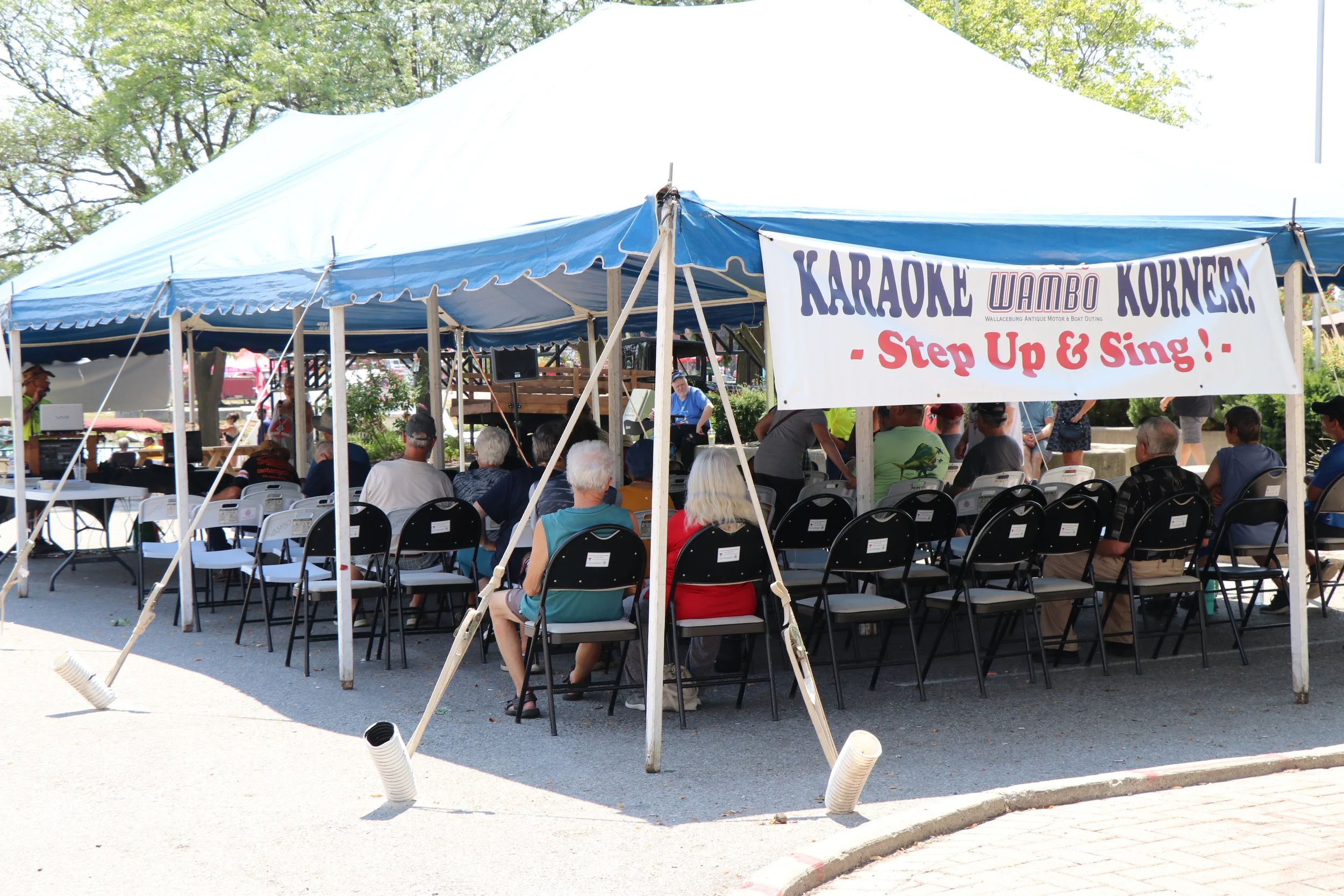 Large blue tent sent up in smaller downtown Wallaceburg parking lot, set up with lots of cars with people seated watching someone at front of tent singing karaoke into a microphone