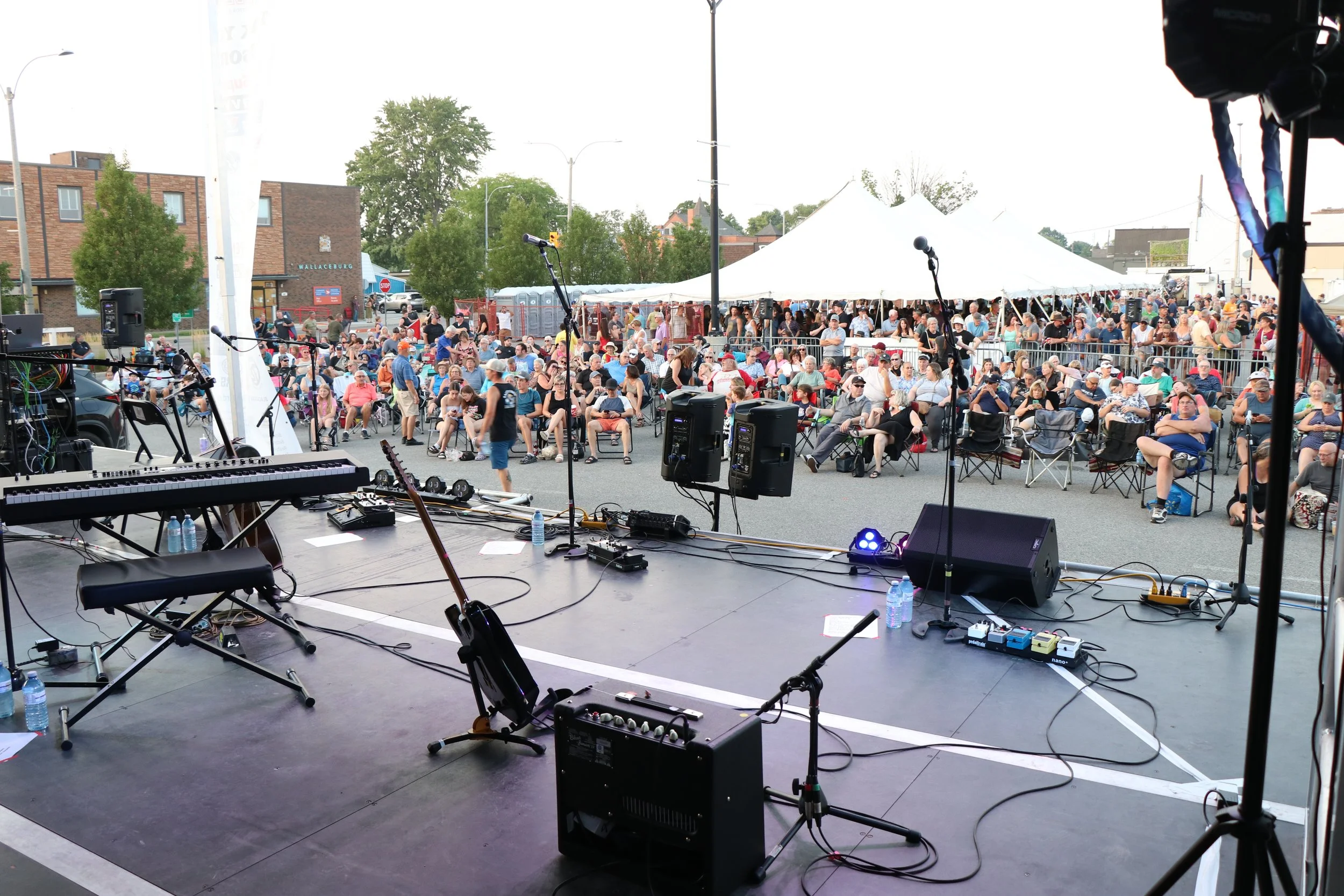 On stage with instruments looking out into large crowd of people seated and in refreshment tent waiting for band to go on