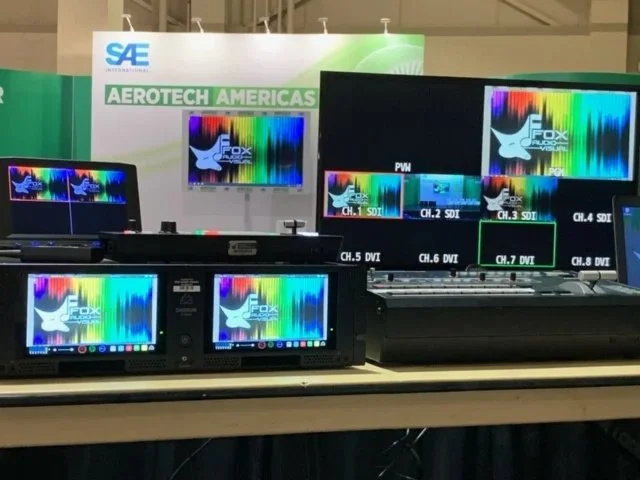 Display monitors and equipment at a trade show booth with a banner labeled 'SAE' and 'Aerotech Americas,' showcasing colorful waveforms and channels.