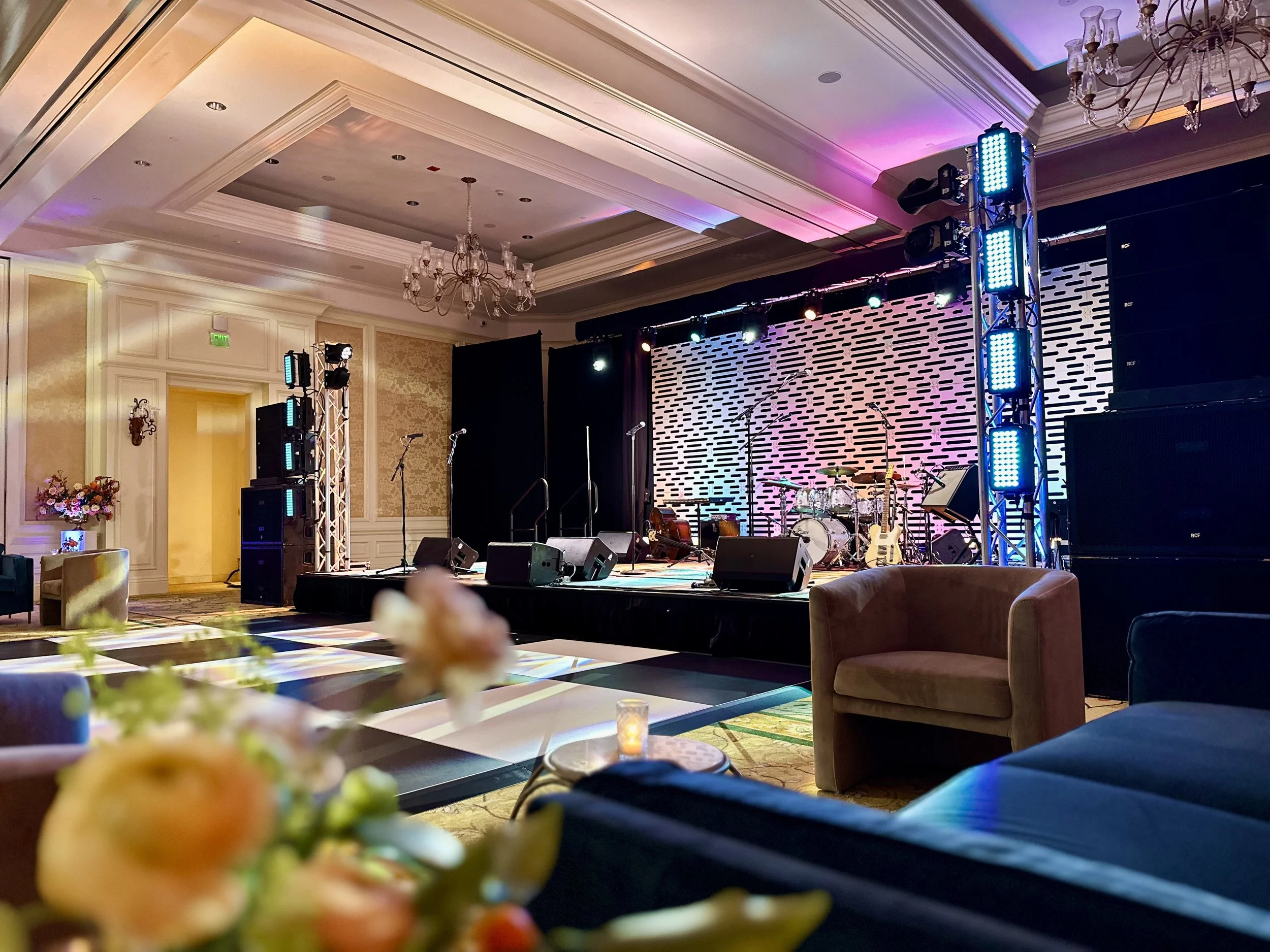 Empty stage with musical instruments and colorful lighting in a banquet hall with seating and floral arrangements.