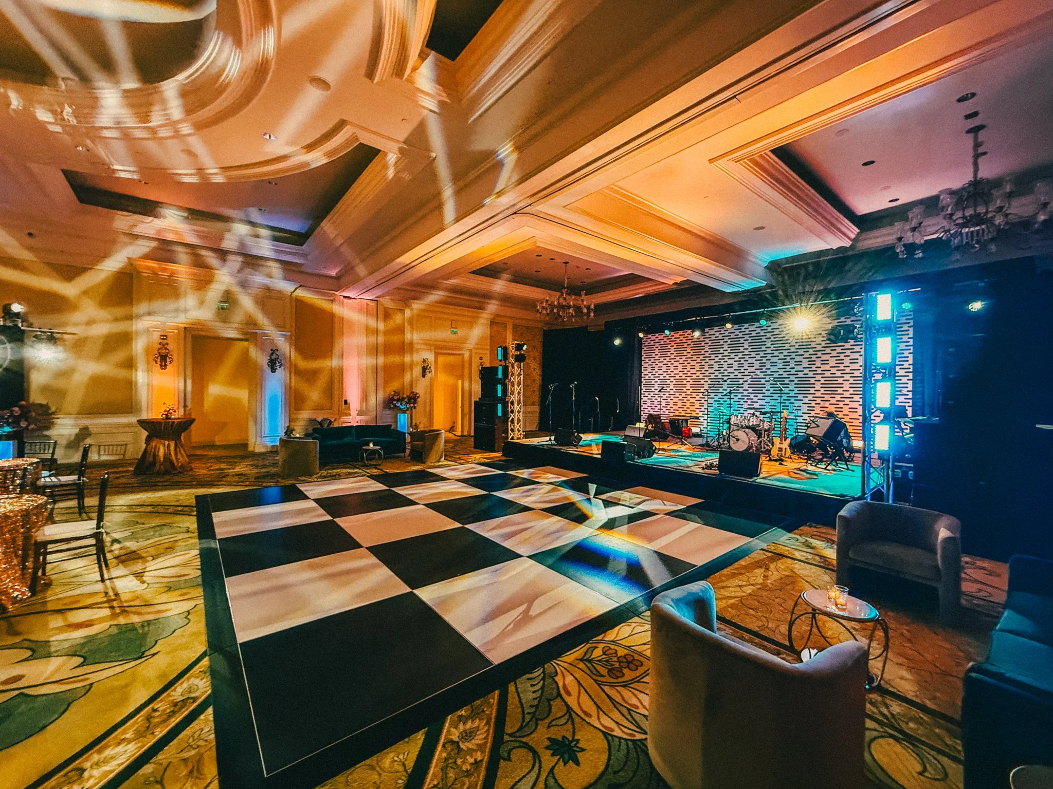 An event hall with a stage, musical instruments, and colorful lighting. The room has a checkered black and white dance floor, elegant ceiling design, and chandeliers. Tables and chairs are arranged around the dance floor.