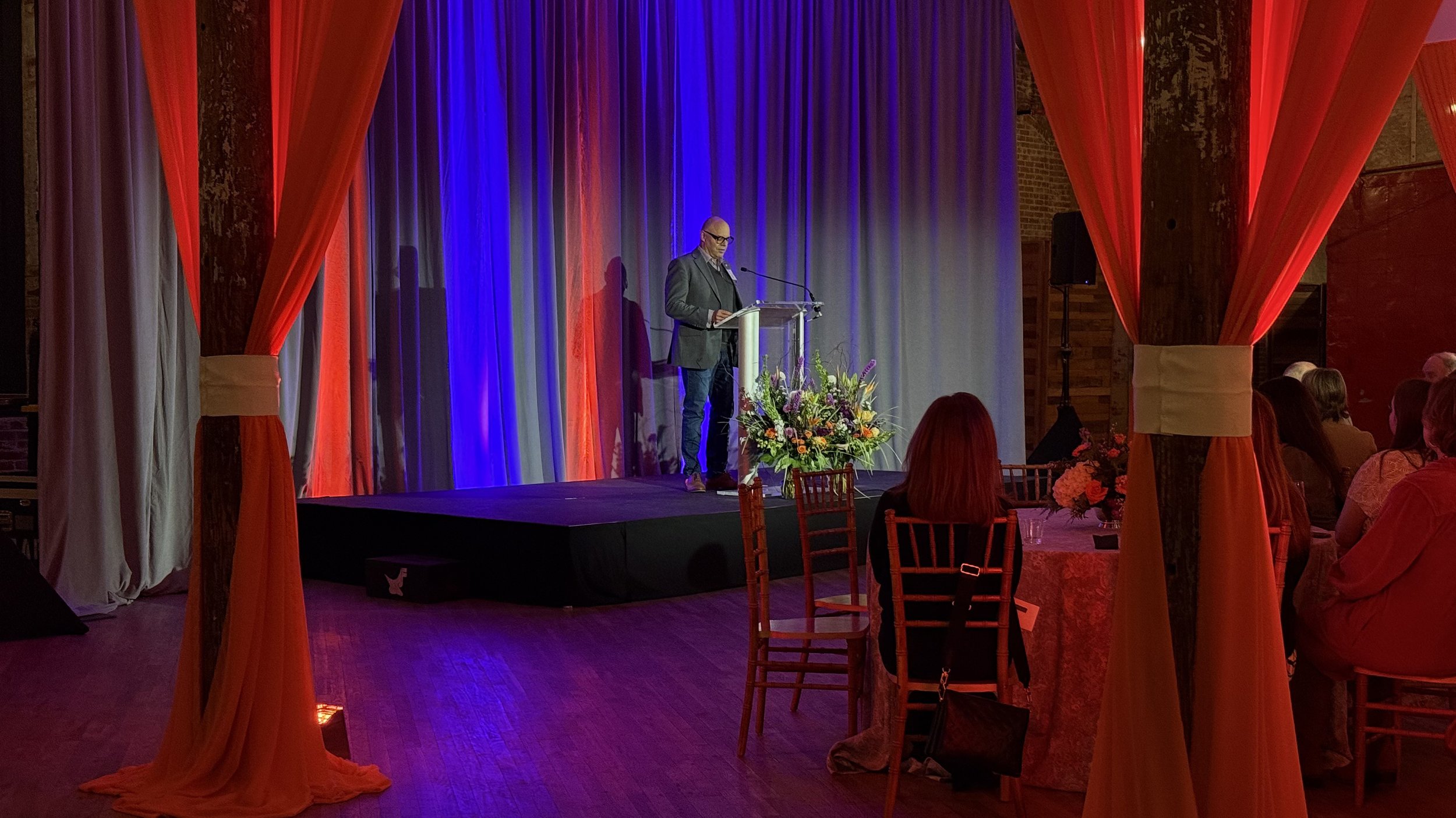 A man stands behind a podium on a stage with a colorful curtain backdrop, speaking during an event with seated attendees at decorated tables.