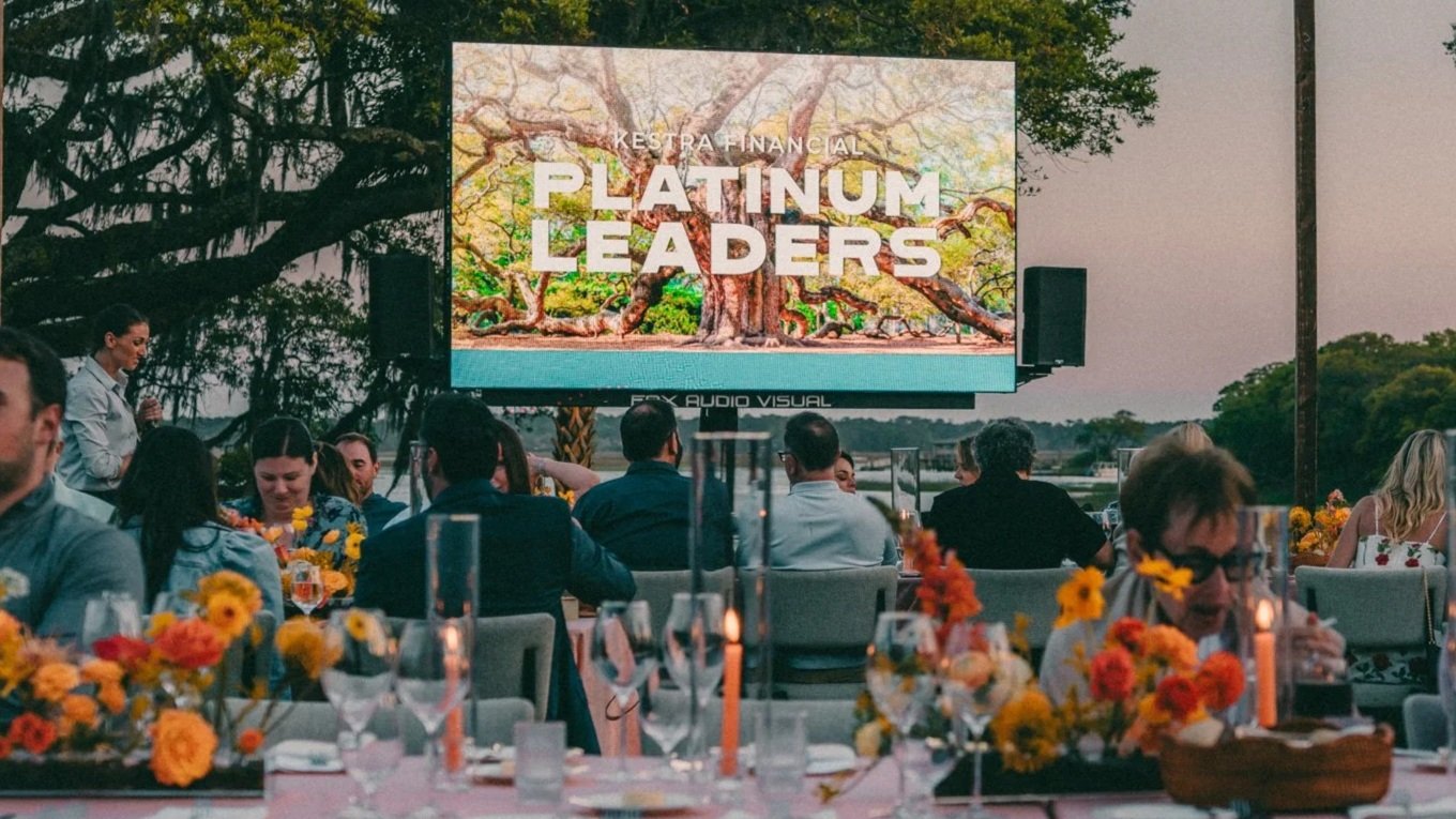 An outdoor event with people seated at tables decorated with flowers and candles, watching a large screen that displays the words 'Kestrel Financial Platinum Leaders' with an image of a tree in the background.