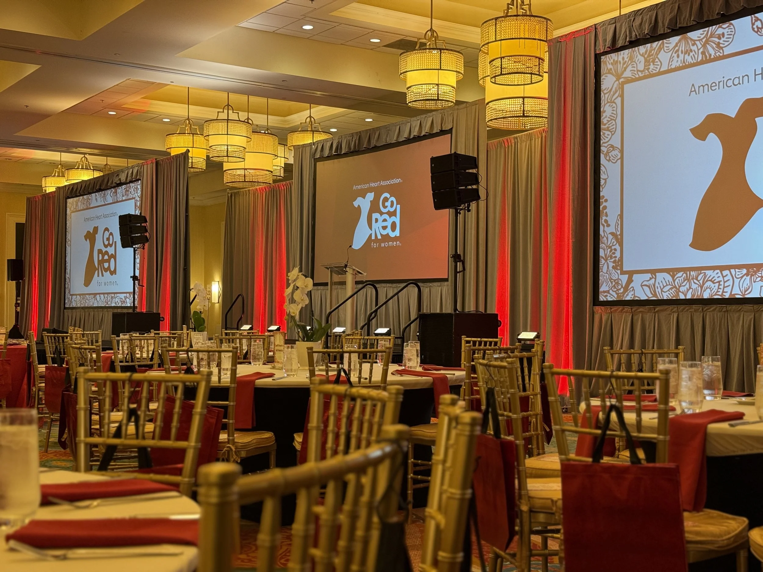 A banquet hall set up with round tables decorated with red and black tablecloths, golden chairs, and centerpieces with white orchids. There are large screens displaying the American Heart Association's Go Red for Women logo, and stage area with podiu