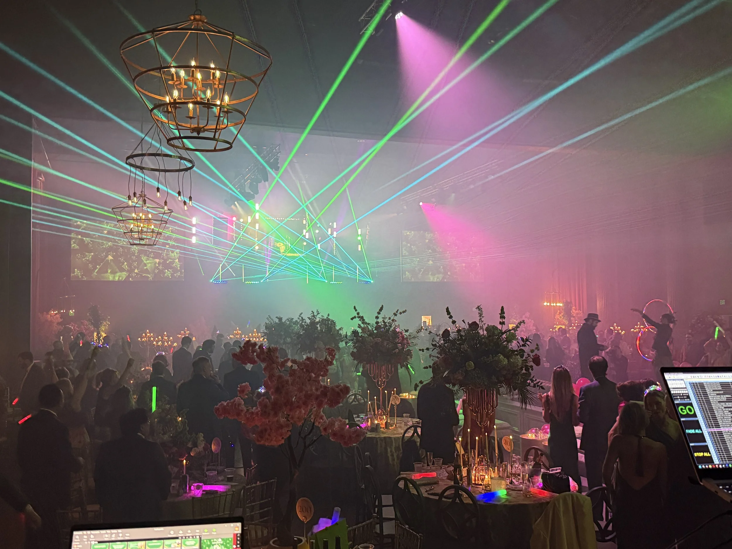 A lively indoor party scene with guests dancing and socializing. The ceiling is decorated with chandeliers, and colorful laser lights illuminate the room, creating a festive atmosphere. There are tables with floral centerpieces and candles, and a DJ 