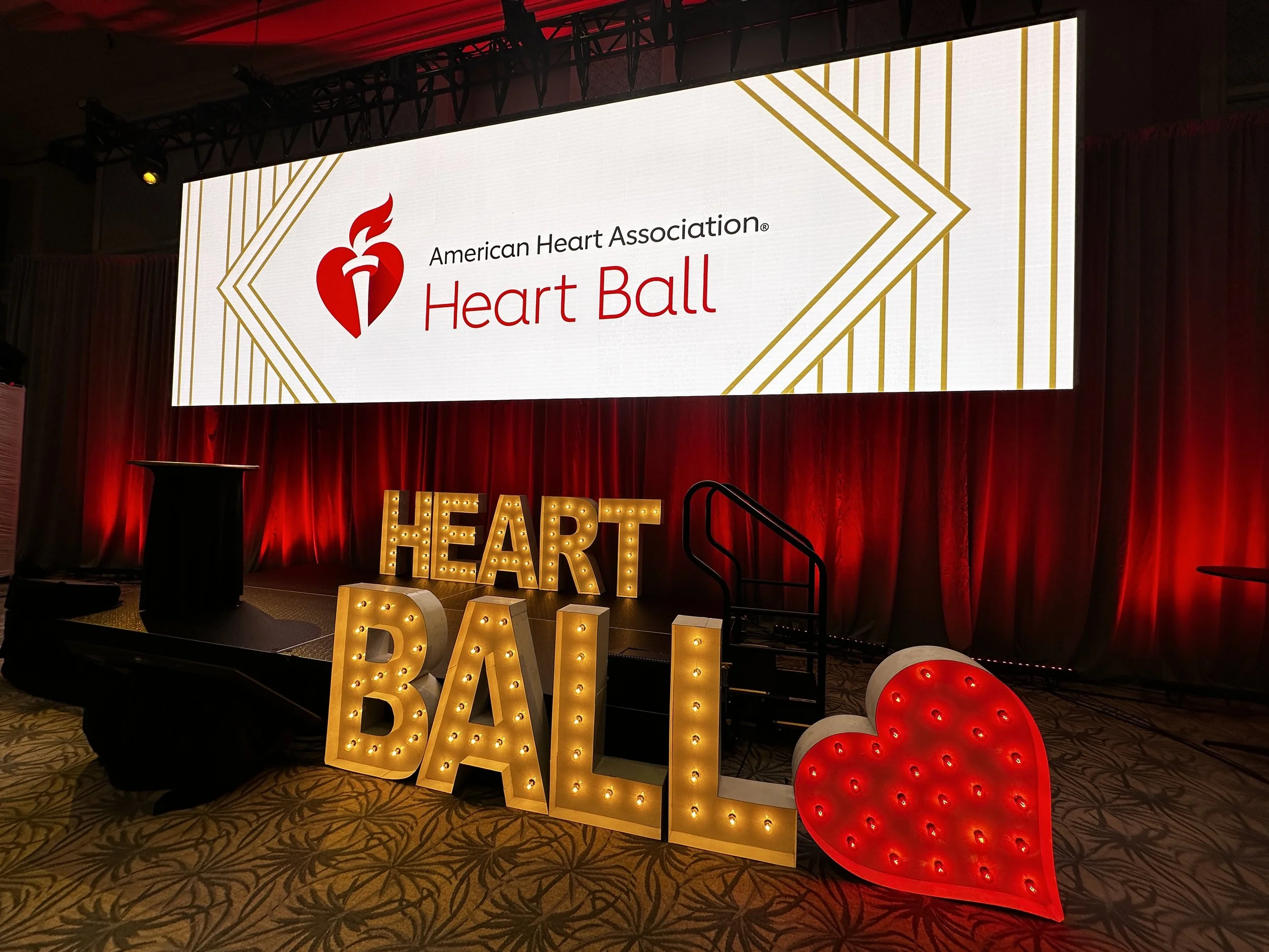 Large display screen showing the American Heart Association Heart Ball logo with a red heart and a flame, and gold geometric patterns. In front, illuminated marquee letters spell 'HEART' and decorative red illuminated hearts are on the floor. The eve