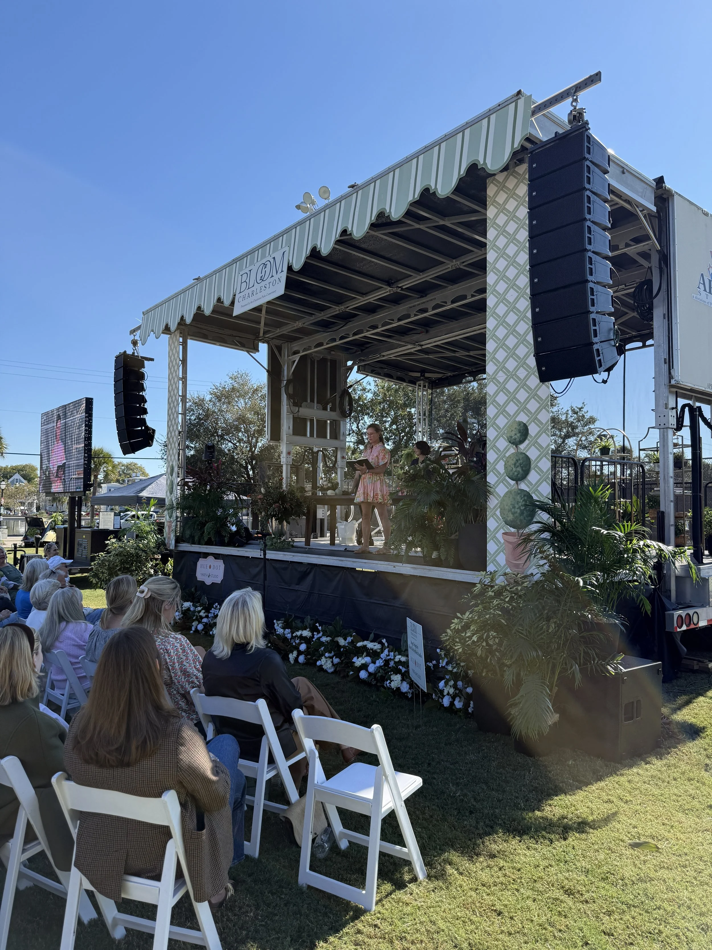 People gathered outdoors watching a woman on a stage with speakers and plants, under a canopy, during daylight