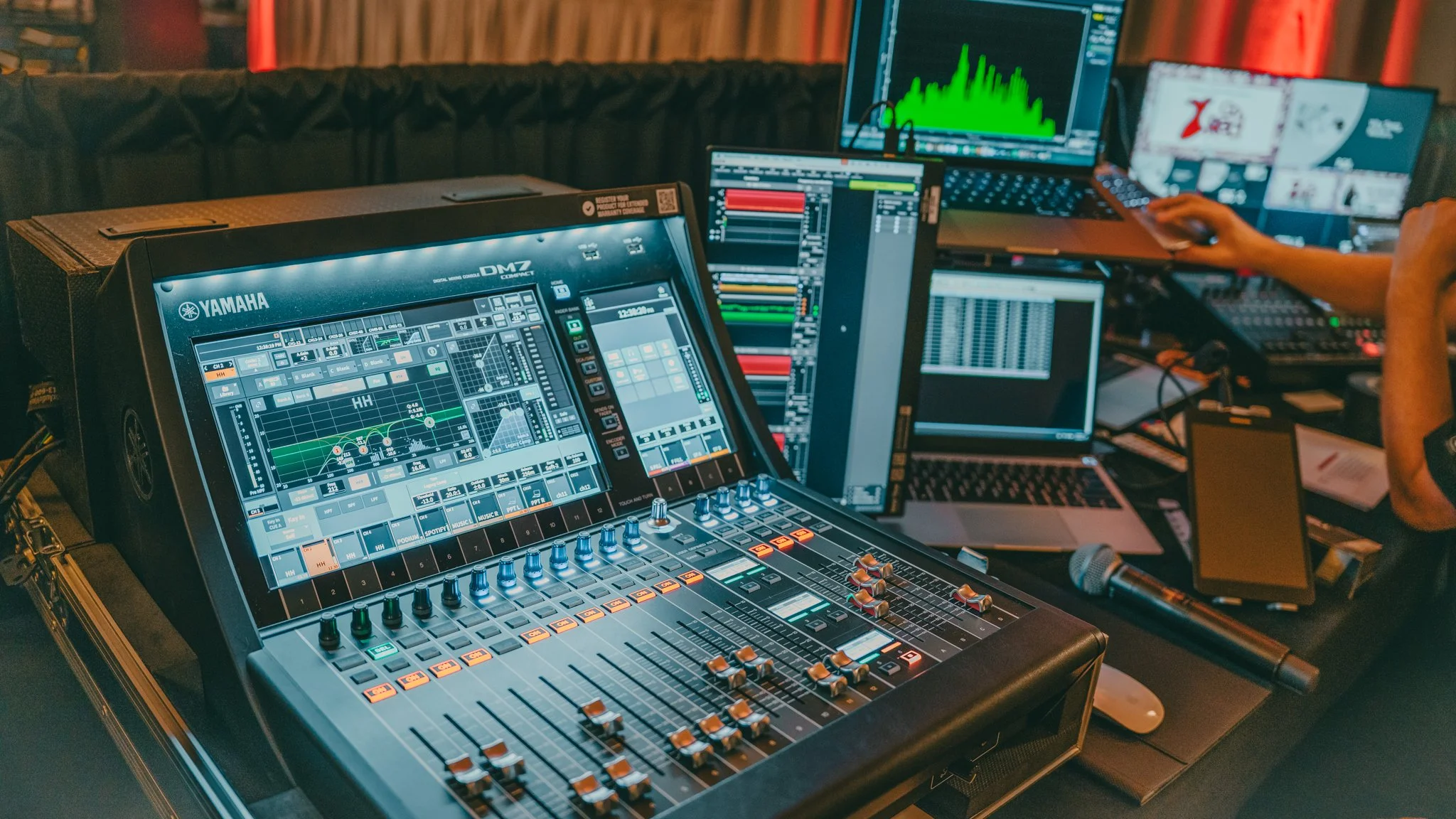 A multi-screen setup with digital audio mixing equipment, laptops, and monitors displaying audio editing software, in a dimly lit room.