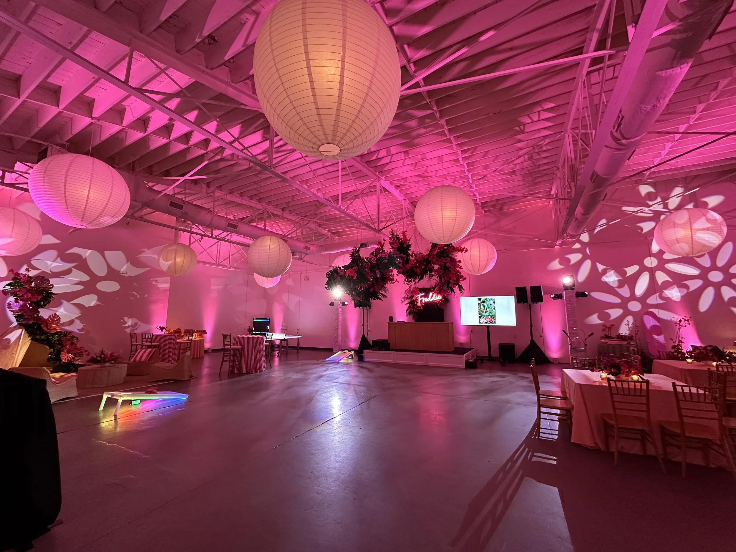 Indoor event space decorated with pink lighting, large paper lanterns hanging from the ceiling, floral arrangements, tables with pink tablecloths, and a stage with a digital screen and sound equipment.