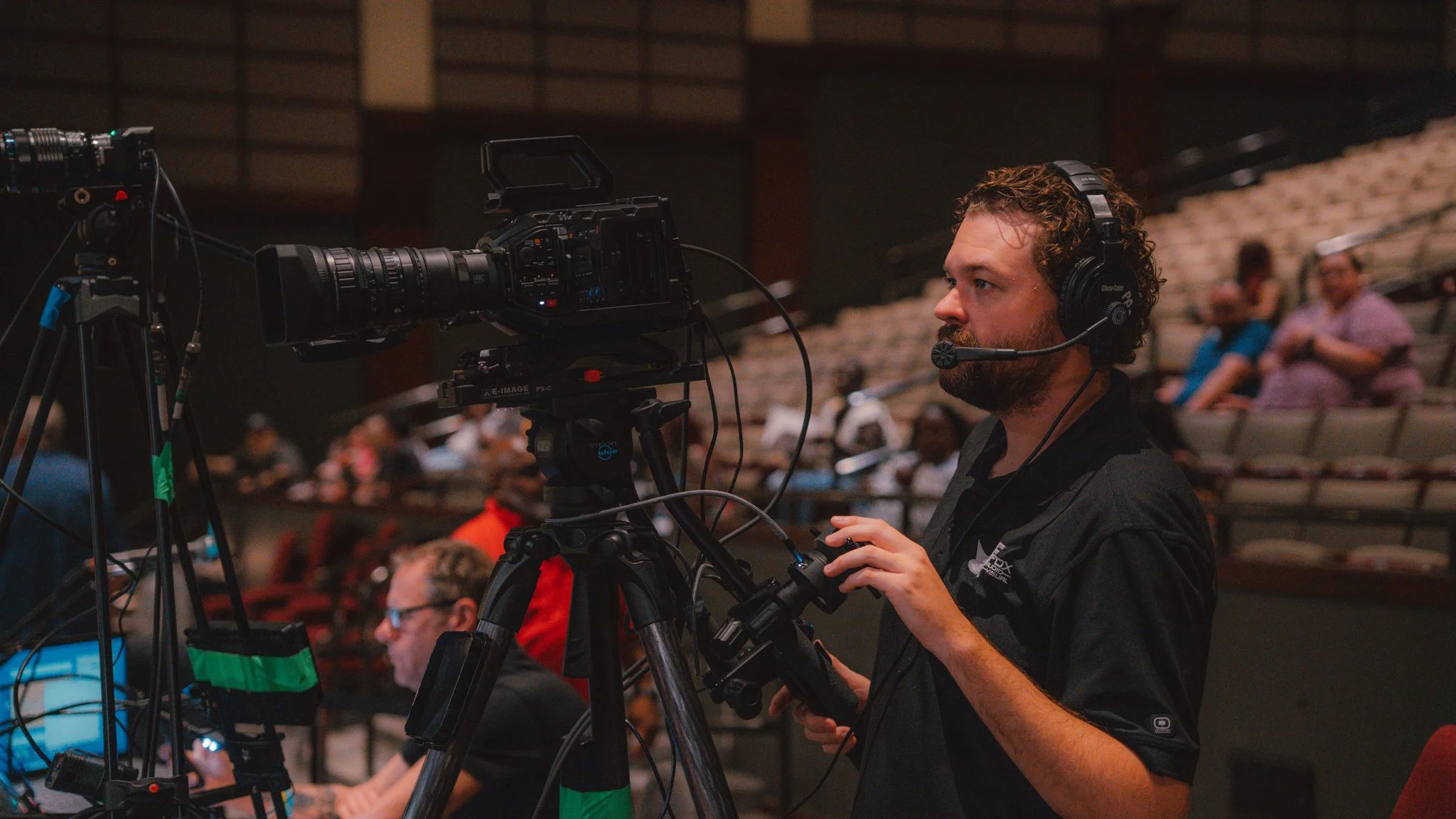 A man with curly hair and a beard wearing a headset operates a professional video camera on a tripod. In the background, several people are seated in an auditorium.