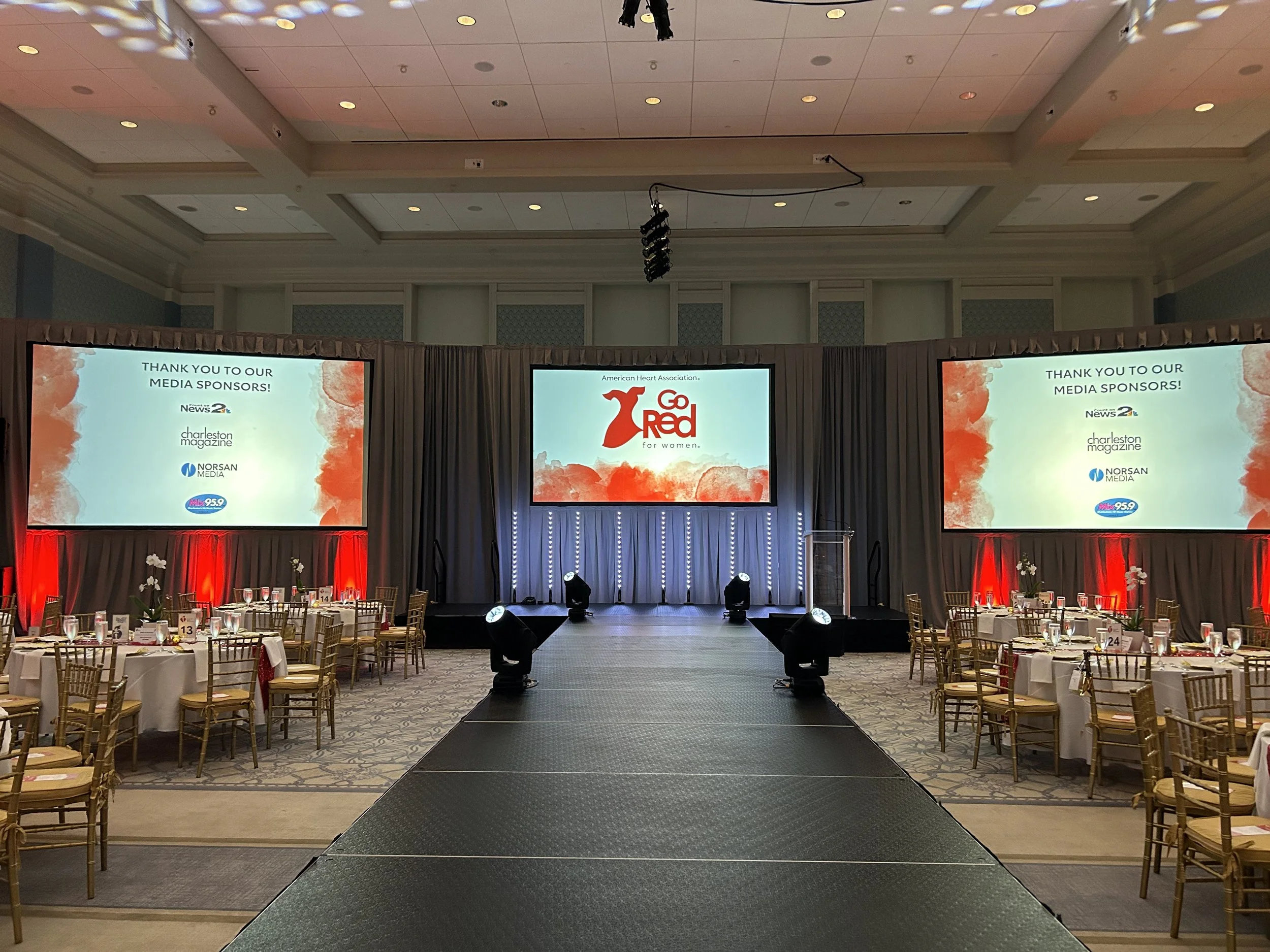 The image shows a banquet hall set up with round tables and chairs, a stage with large screens displaying the American Heart Association's Go Red for Women campaign, and decorated with red and white accents. There are lighting and audio equipment han