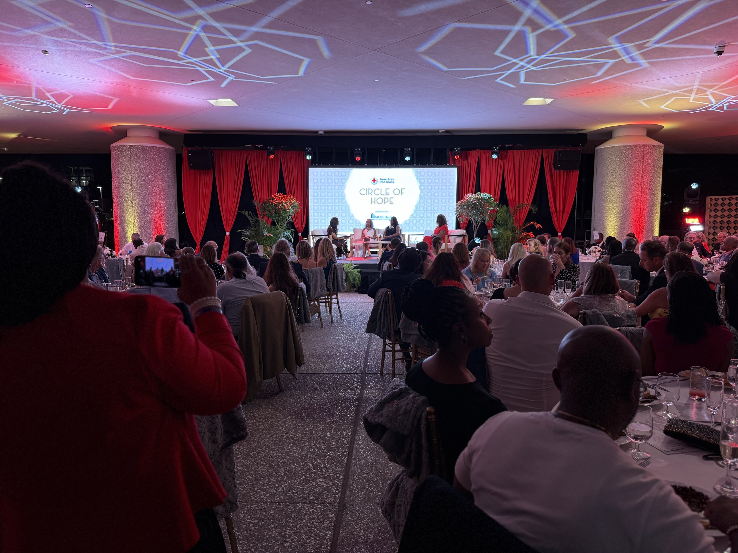 An indoor event with a stage in the background featuring a large screen displaying 'Circle of Hope' and 'American Red Cross'. The stage is decorated with red curtains and large plants. The audience is seated at round tables, with some people taking p