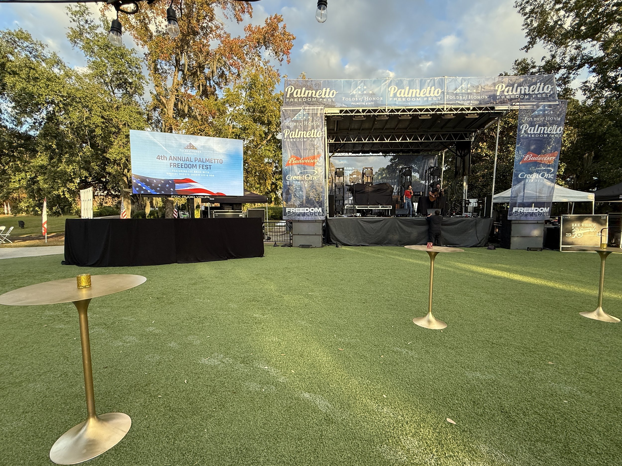 An outdoor stage setup at the Palmetto Freedom Fest with promotional banners, two musicians, and a large screen displaying event information, surrounded by green trees and a clear sky.