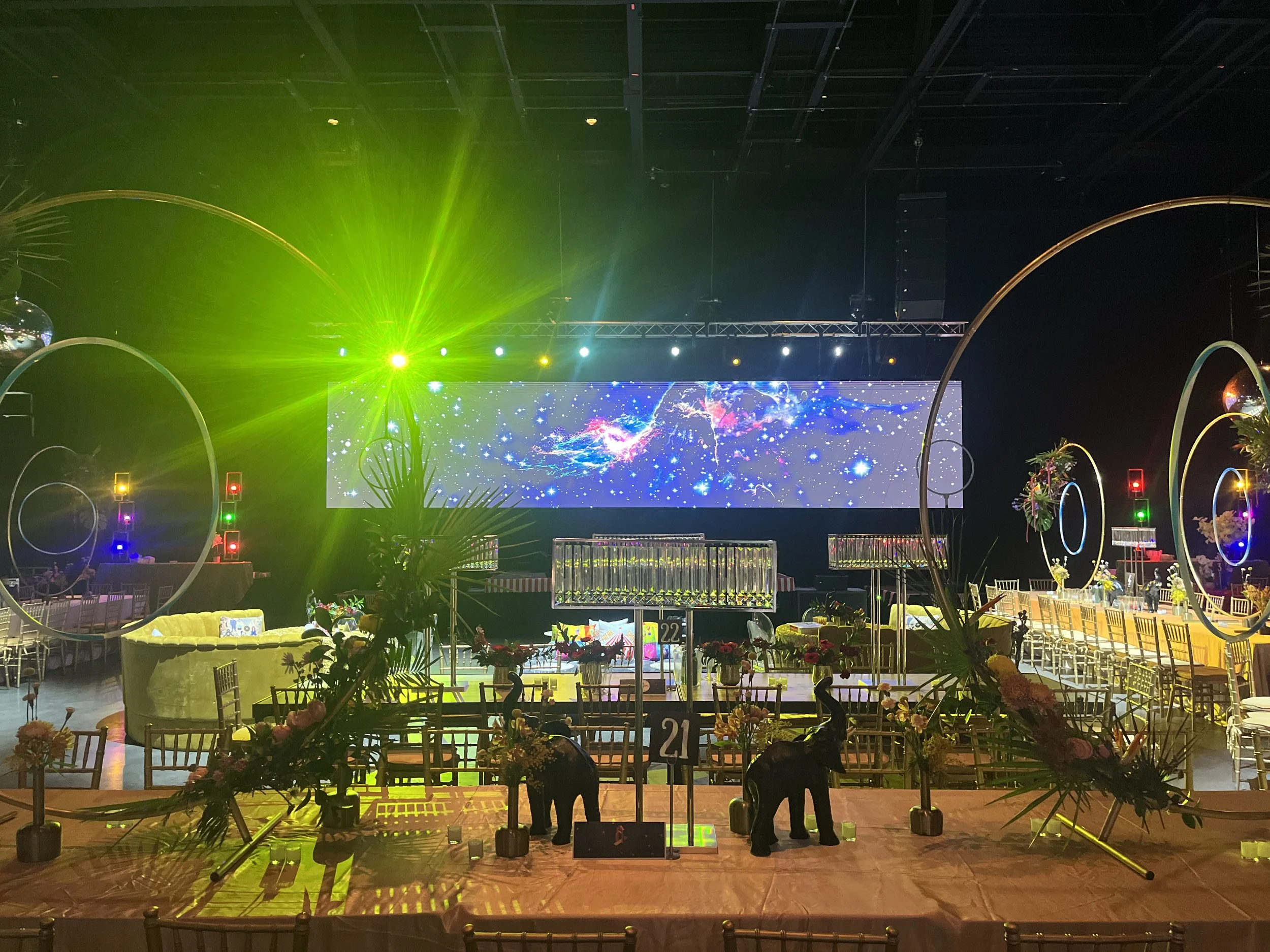 A stage with colorful lights and a large digital screen displaying a cosmic scene with stars and nebulae. There are chairs and tables set up in front of the stage, decorated with flowers, and a decorated arch on the sides of the stage. The ceiling is