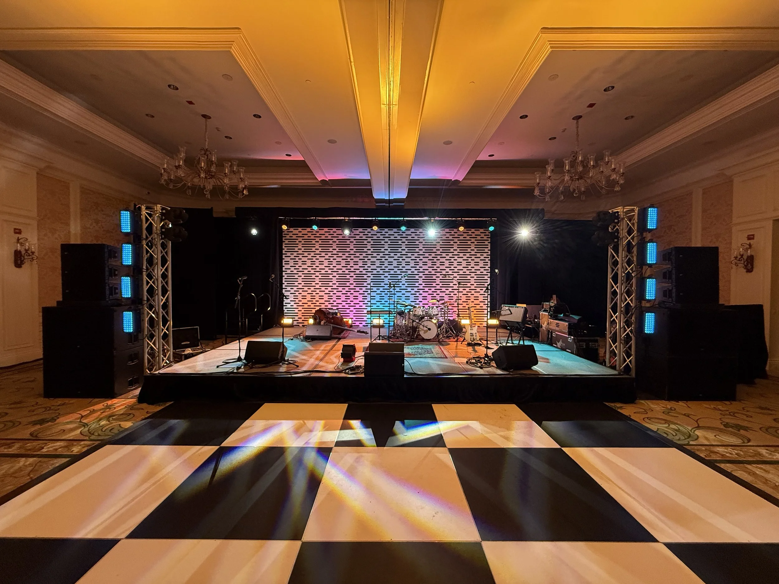 Empty stage set up for a live music performance with various musical instruments and speakers, illuminated with colorful stage lights and a patterned backdrop.