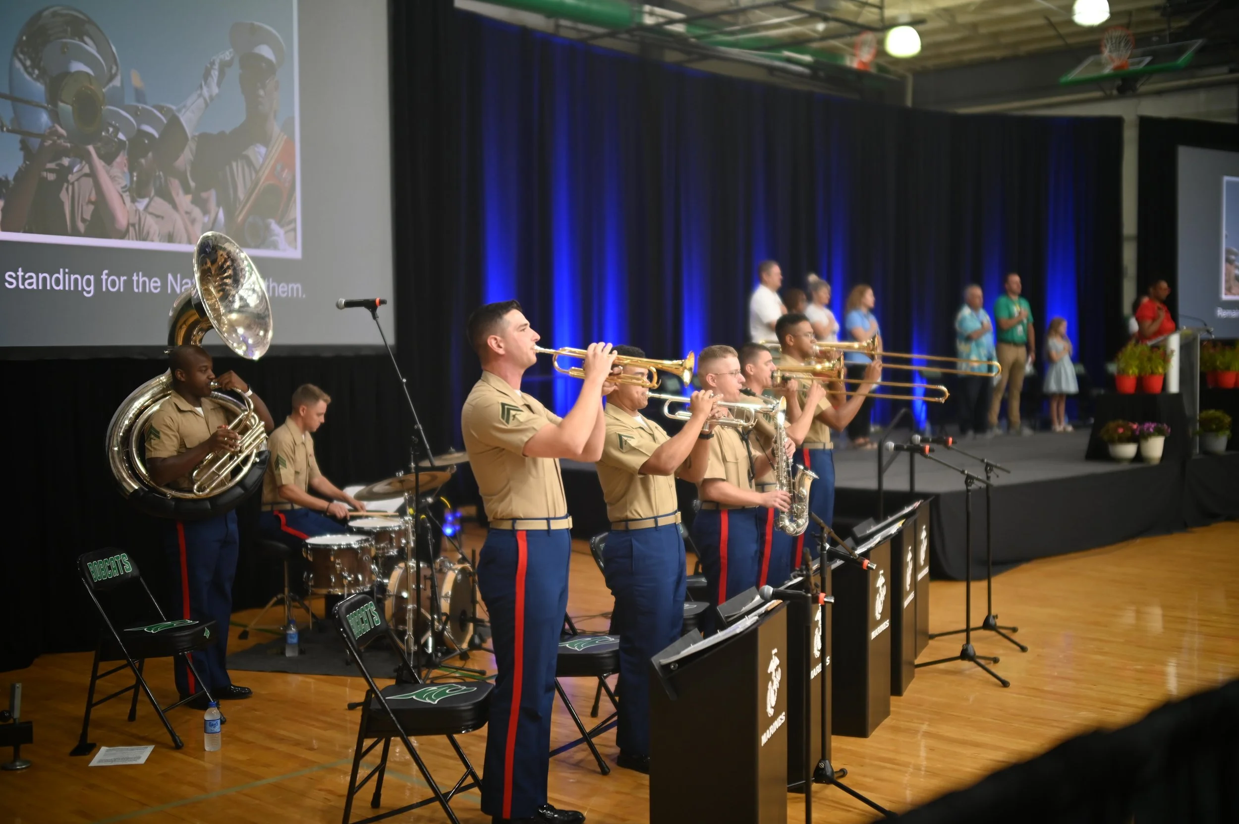 Military band performing on stage with musicians playing trumpets, saxophones, drums, and tuba, with a large screen displaying an image of soldiers and text in the background, and a group of people standing on the side of the stage.
