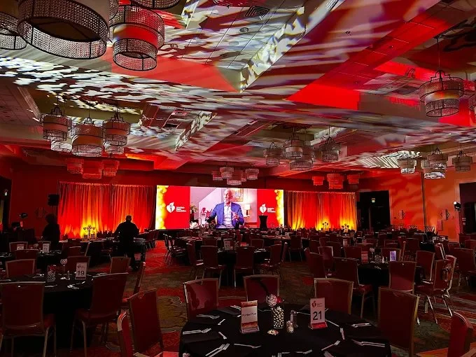 An event space decorated with red and orange lighting, featuring round tables with black tablecloths and chairs, a large stage with a speaker in a suit, and a big screen displaying the speaker's image, with ceiling lights and patterned ceiling reflec