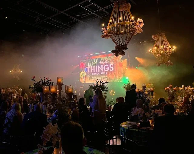 Audience at a formal event with elegant chandeliers, large floral arrangements, and a colorful stage display reading 'Wild Things Annual Gala' in a dimly lit setting.