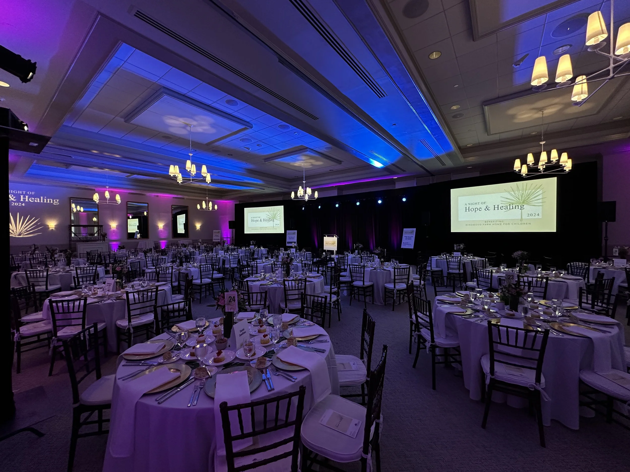 Indoor banquet hall decorated for an event with tables set with white tablecloths, wine glasses, and small desserts, illuminated with purple and blue lighting, and large screens displaying event information on stage.