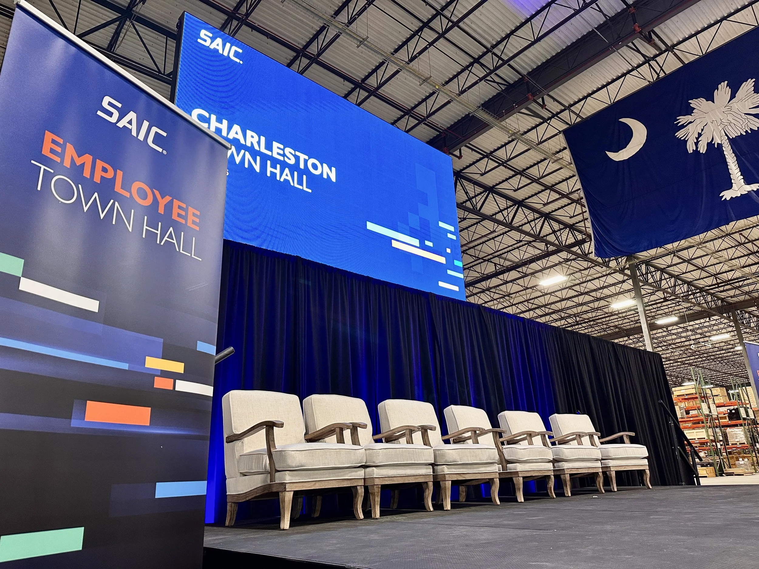An indoor stage with four beige chairs with dark wood armrests and legs. Behind the chairs, there is a blue stage backdrop with the SAIC logo. To the left, a tall banner displays the SAIC logo and the words "Employee Town Hall." To the right, there's