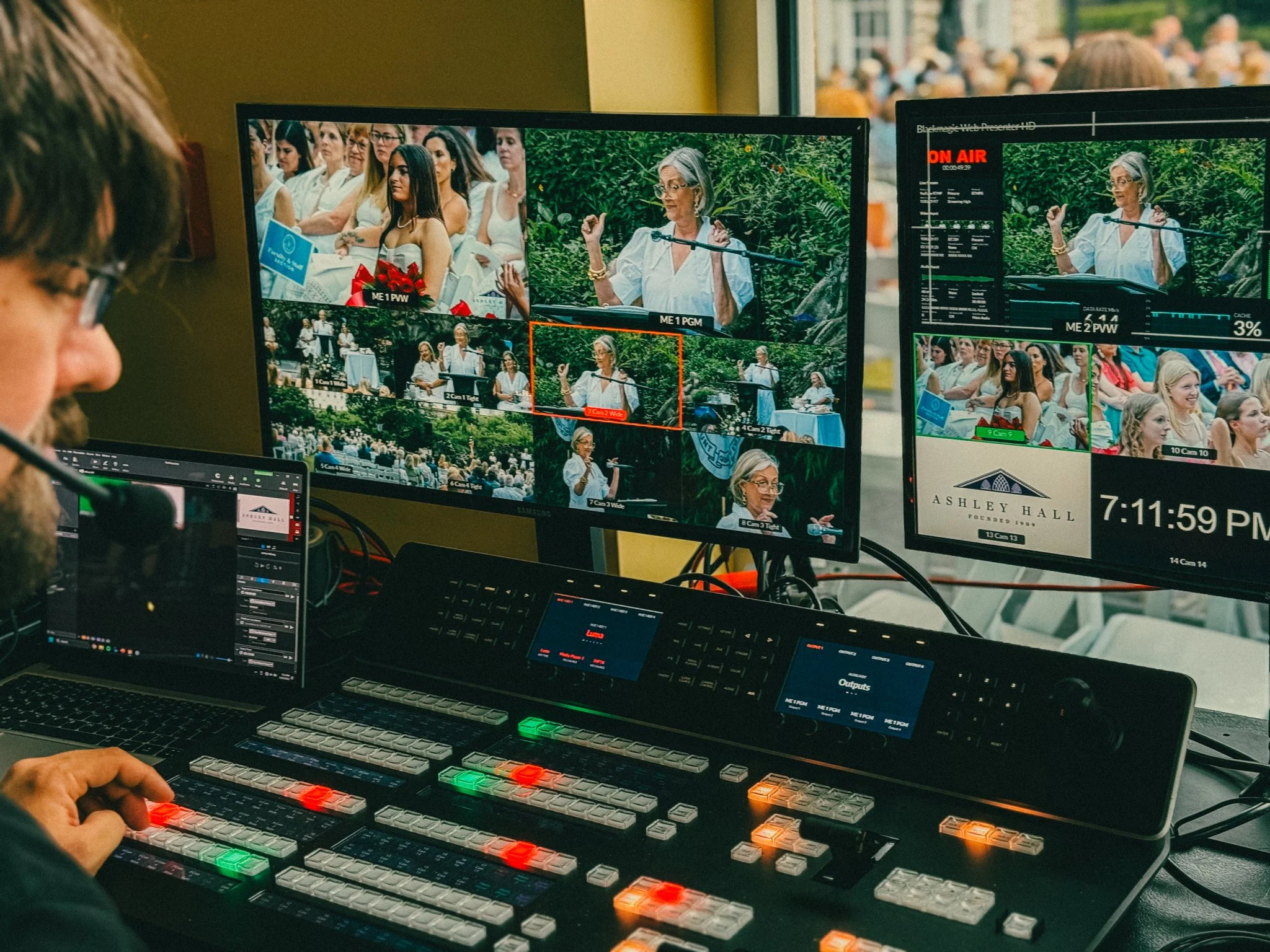 Video editing setup with multiple monitors displaying a woman speaking at an outdoor event, with audience in the background, and a control panel with colorful buttons in the foreground.