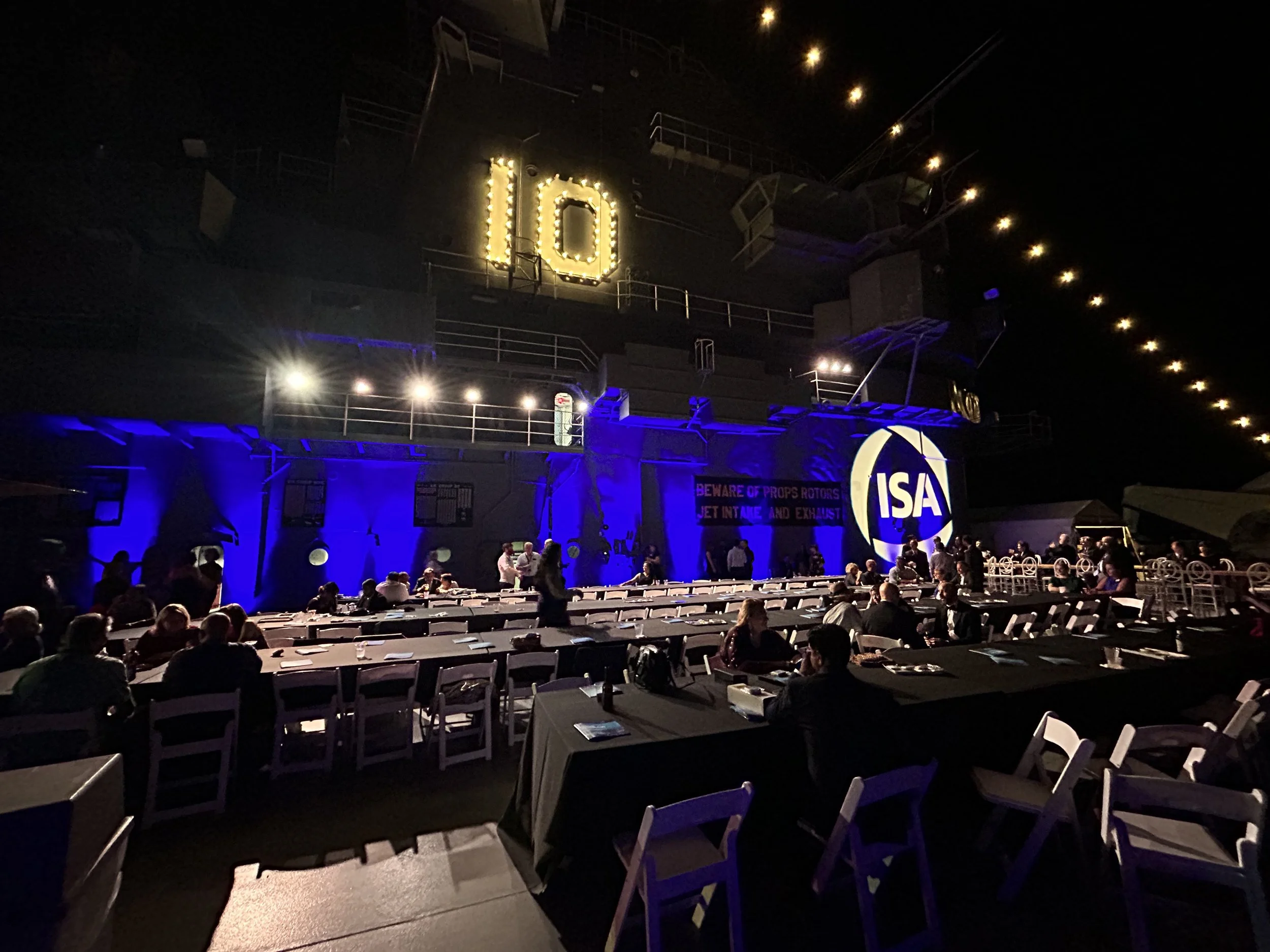 Nighttime scene of an auditorium with white tables and chairs, illuminated by blue and yellow lighting, with a large illuminated sign displaying the number 10 and the NASA logo in the background.