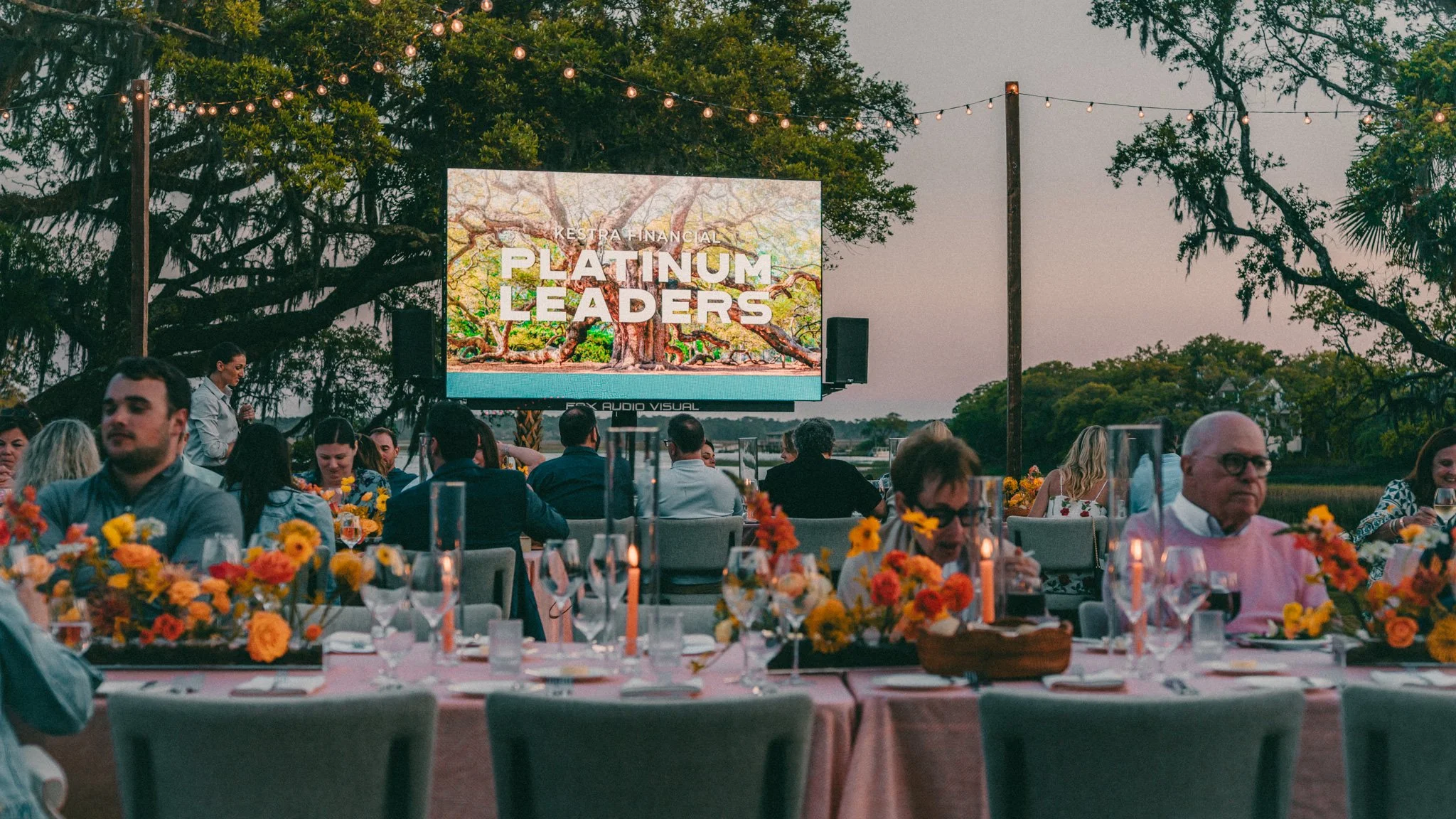 An outdoor event with a large screen displaying 'Platinum Leaders'. People are seated at tables decorated with flowers, and string lights hang overhead. The scene is set in the evening with trees in the background.