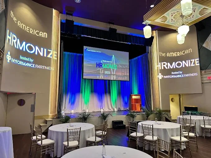 Event stage set up with large screen displaying 'Welcome to HRmonize 2023', surrounded by curtains illuminated with blue and green lights, with tables and chairs arranged in front, and large screens on either side displaying event branding.