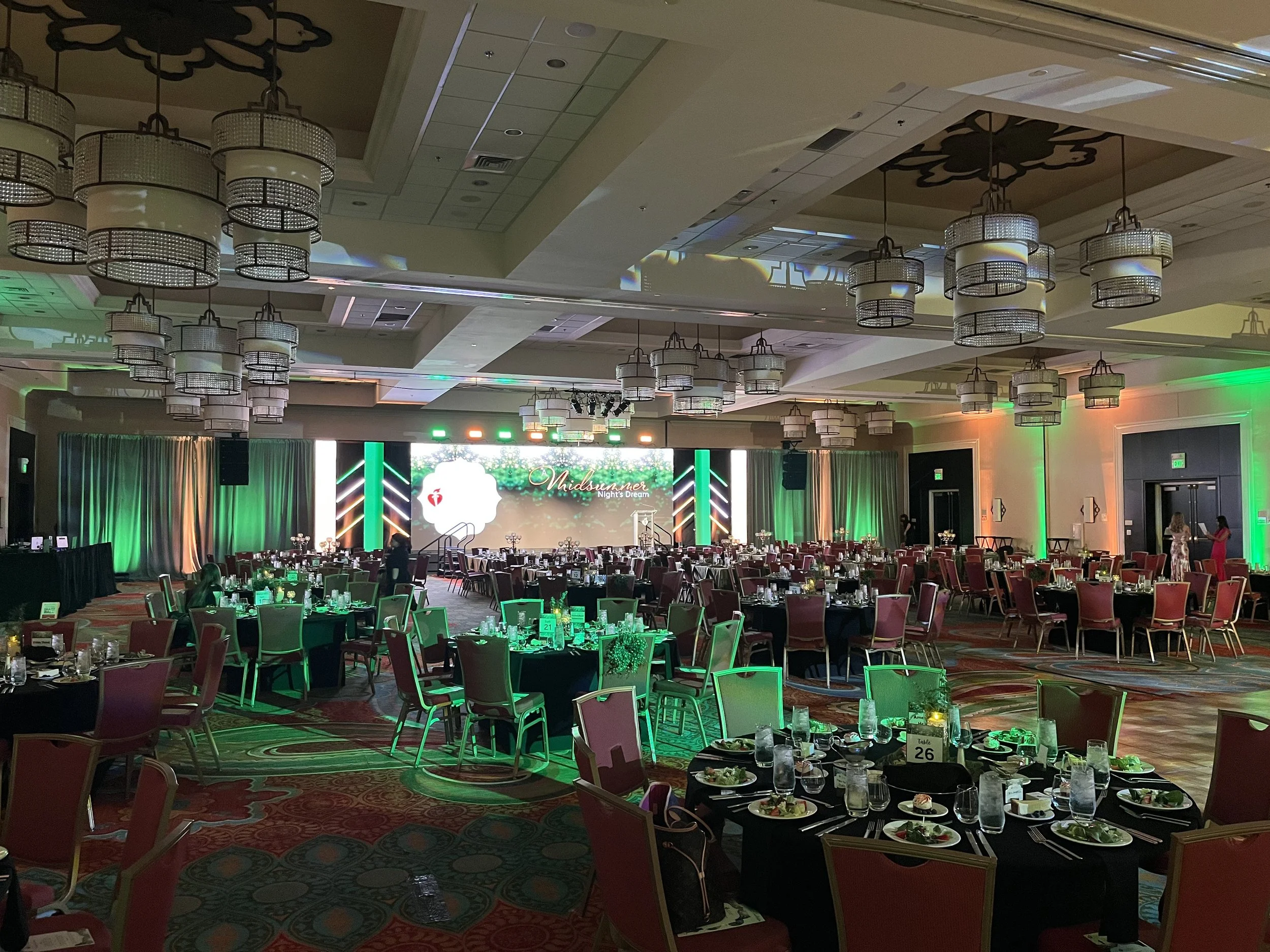 A large banquet hall decorated for an event with round tables covered in black tablecloths, set with plates, glasses, and silverware, and illuminated with green and warm lighting. The background features a stage with a large LED screen displaying the