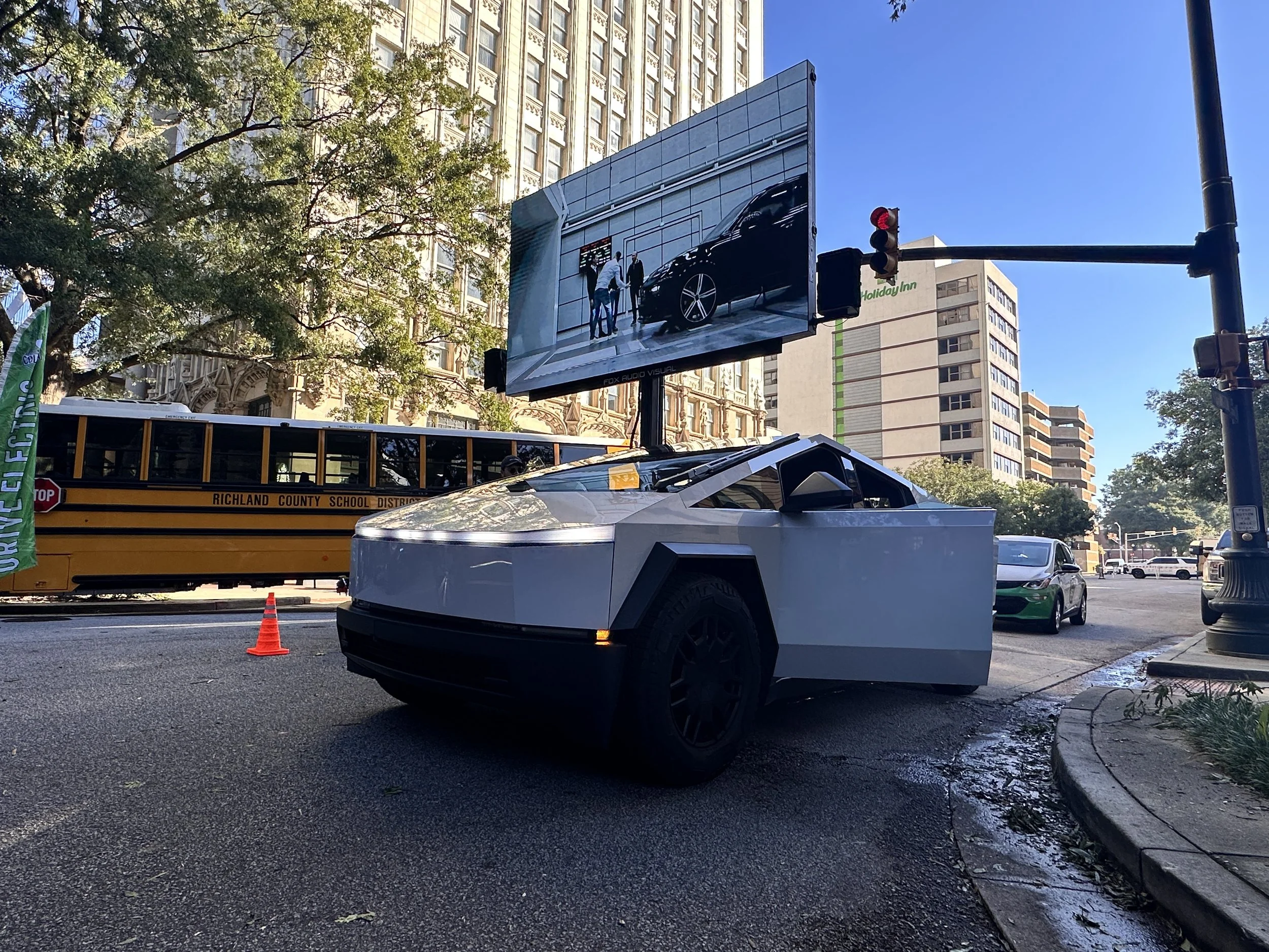 A city street scene with a modern robotic vehicle, a yellow school bus labeled 'Richland County School District,' a traffic cone, and a digital billboard displaying a reflection of a car and two people. Tall buildings and trees are visible in the bac