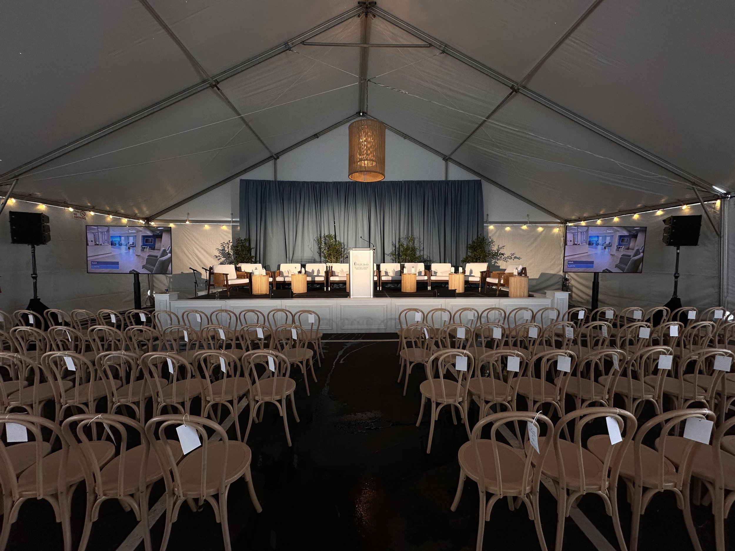 Empty indoor event tent with chairs arranged facing a stage with a podium, blue curtains, potted plants, and two large screens on either side.