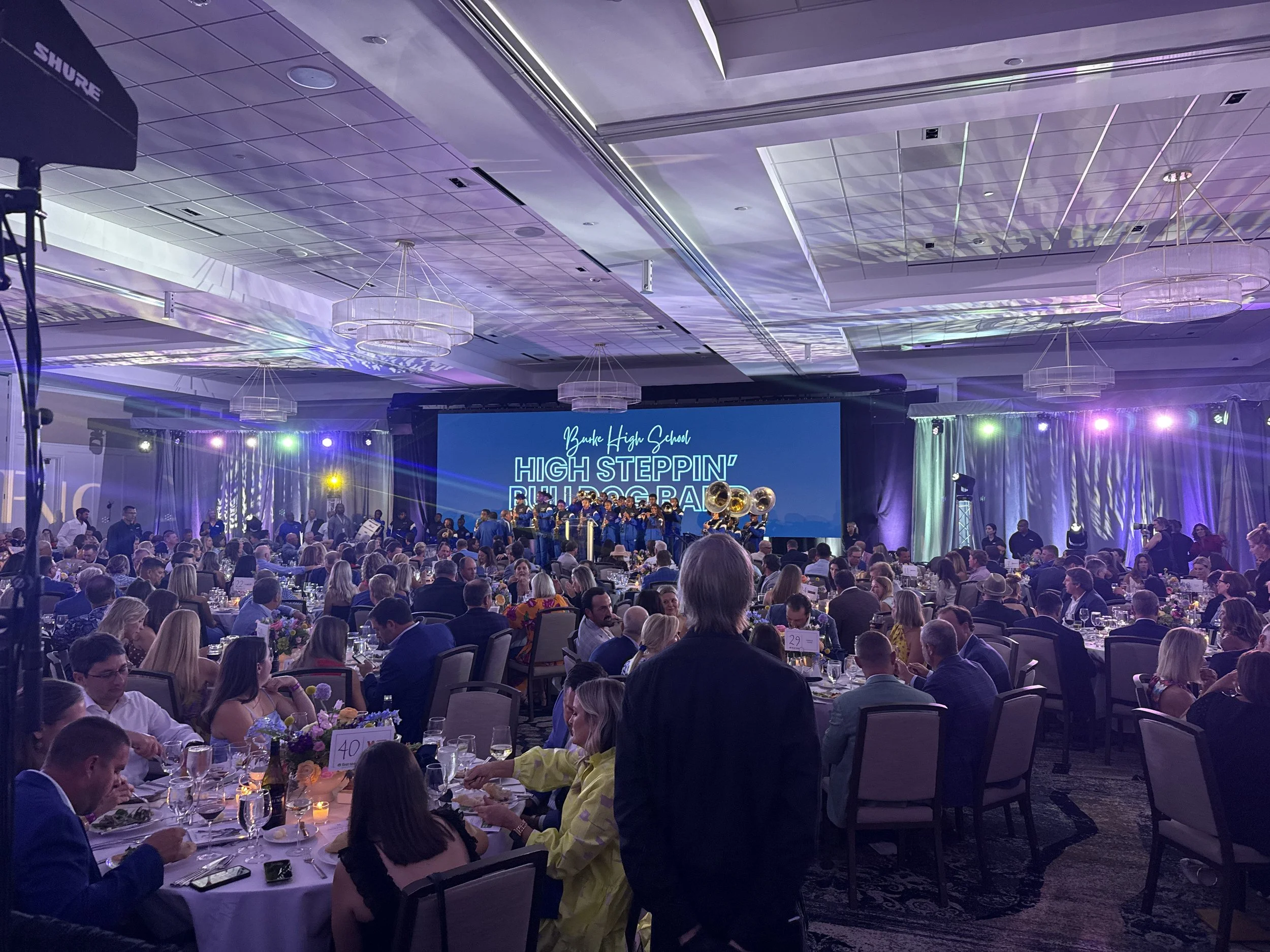 A large indoor banquet hall filled with people seated at round tables, watching a performance on stage. The stage features a sign that says 'High Steppin' Band' and a band performing with musical instruments. The room is decorated with chandeliers an