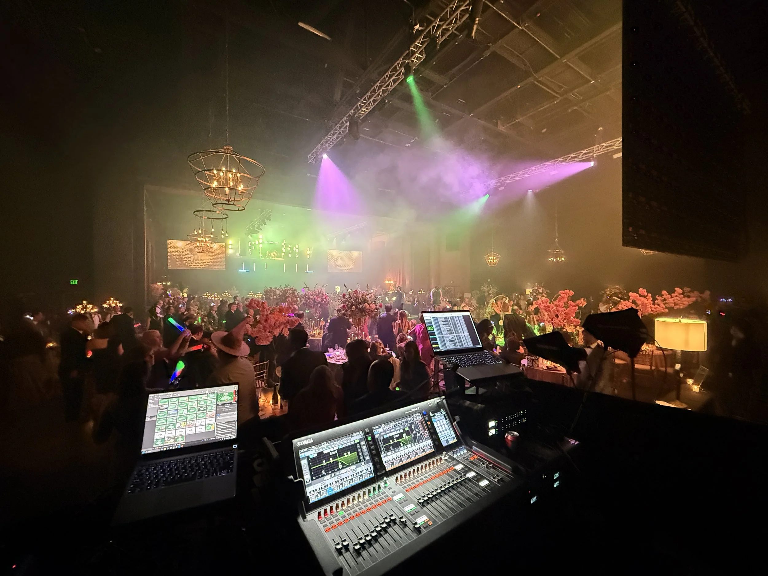 A crowded indoor event with stage lighting, floral arrangements, and a DJ setup with electronic equipment overlooking the dance floor.