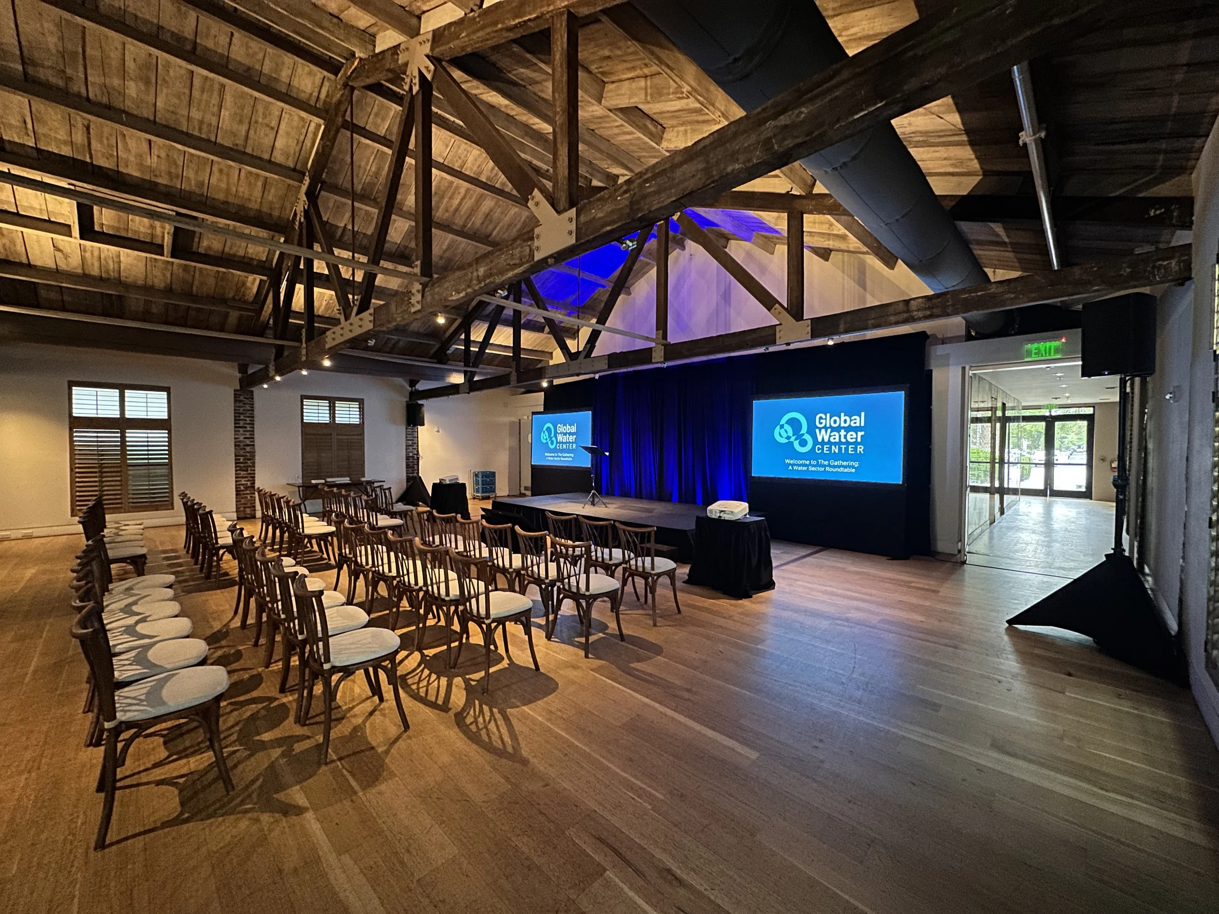 An indoor event space with rows of chairs facing a stage with two large screens displaying 'Global Water CENTER'. The space has wooden flooring, exposed wooden beams on the ceiling, and a door with an 'Exit' sign to the right.