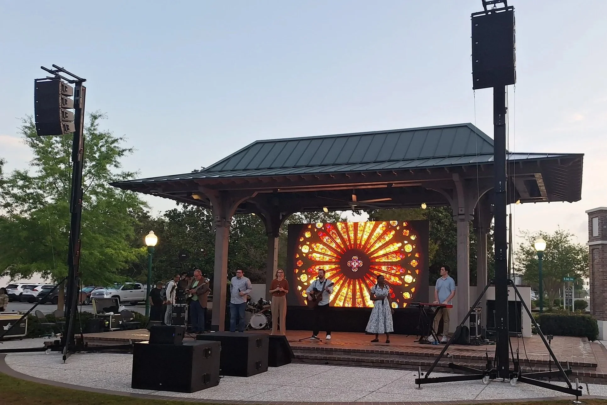 An outdoor stage with a band performing, featuring a large digital backdrop displaying a colorful sunflower design. There are multiple musicians, including a guitarist and singers, with speakers and sound equipment surrounding the stage. People are gathered around watching the performance, and the setting appears to be in a park during the early evening.