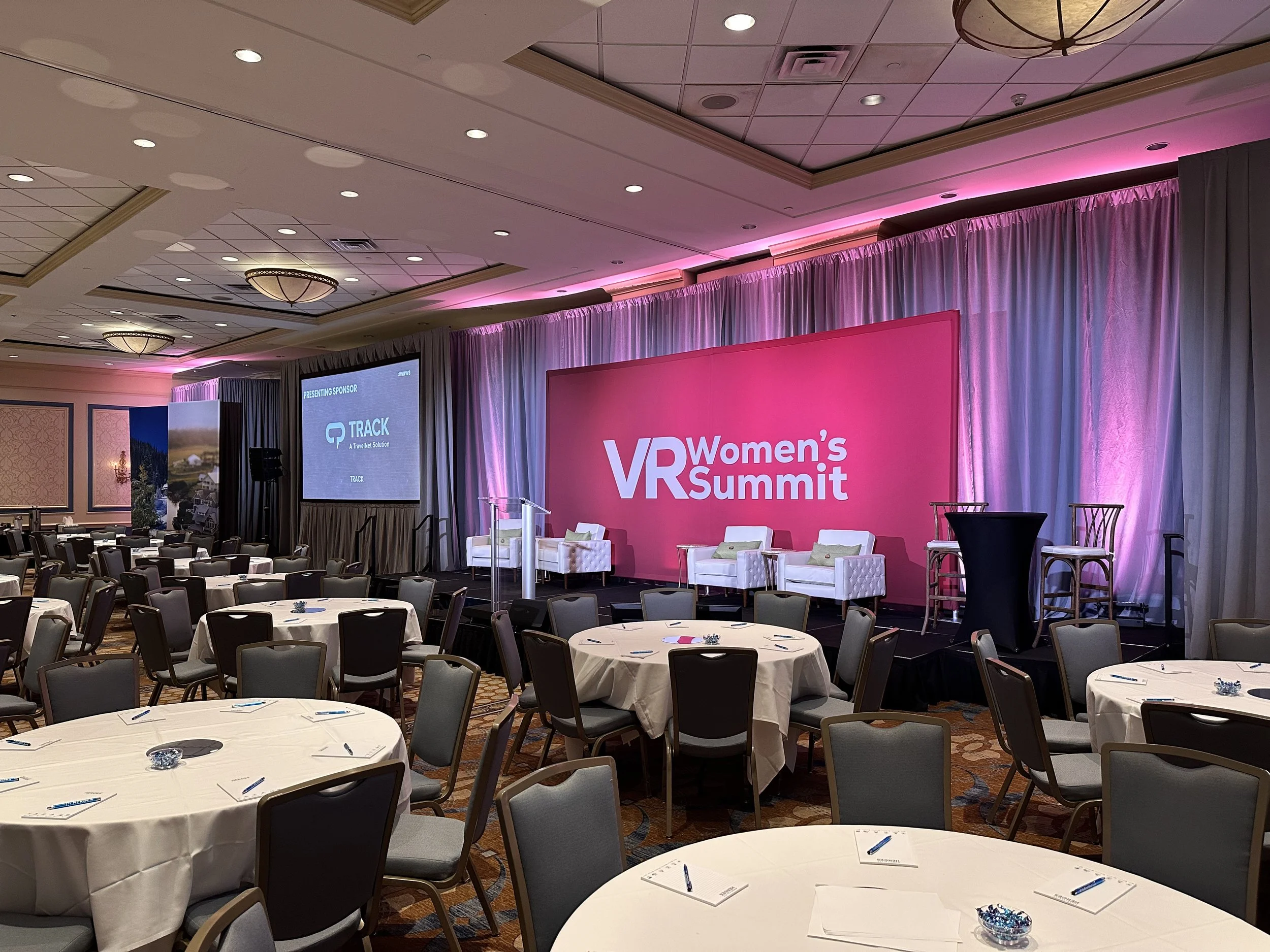 Conference room with round tables, chairs, a stage with a pink backdrop, and projection screens at the VR Women’s Summit.