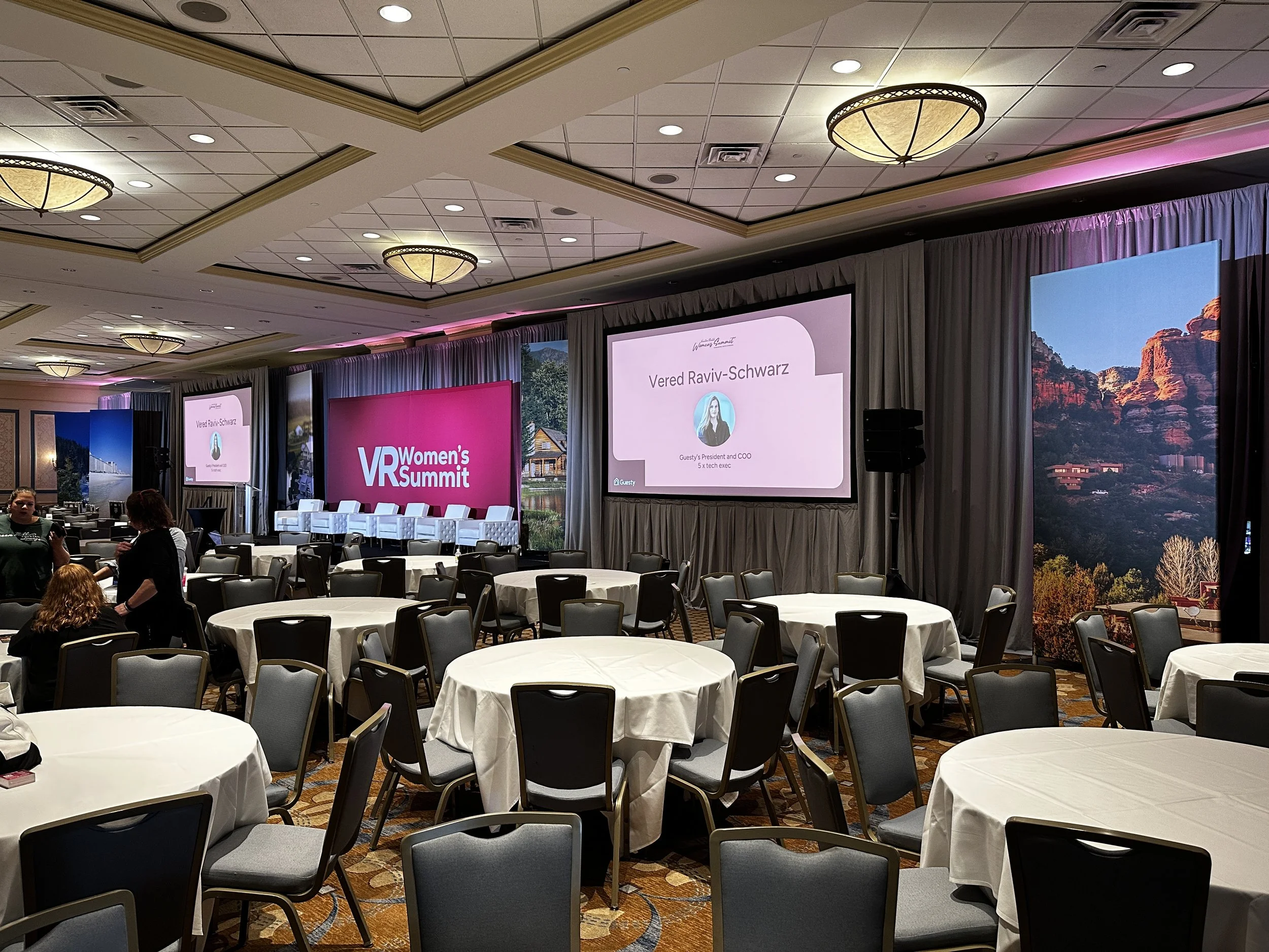 Conference hall with round tables covered in white tablecloths, chairs set around, and a stage with multiple screens displaying the event name 'VR Women's Summit' and a speaker's slide. The hall has a decorated ceiling with chandeliers and scenic bac