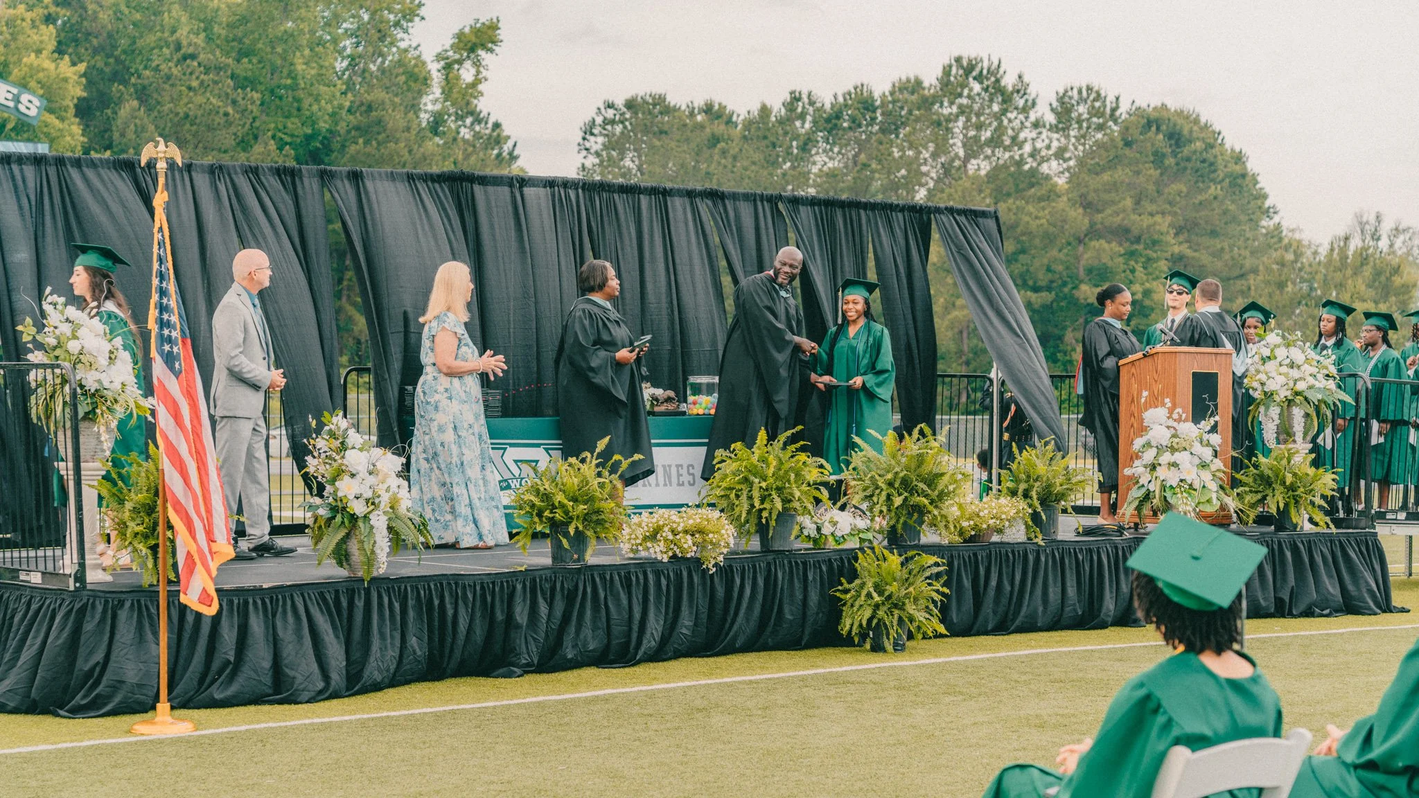 Graduation ceremony on an outdoor stage with students in green caps and gowns. A black male graduate is shaking hands with a man, possibly an administrator, as she receives her diploma. Several other graduates are waiting in line on stage, and others