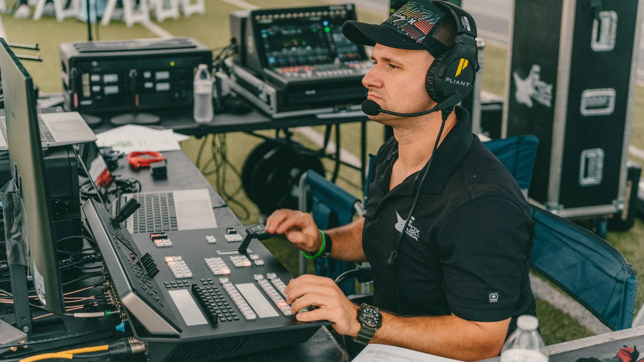 A man wearing a Pliant branded cap and headset is operating audio equipment at an outdoor event. He is focused on a mixing console with various controls and devices on a table.