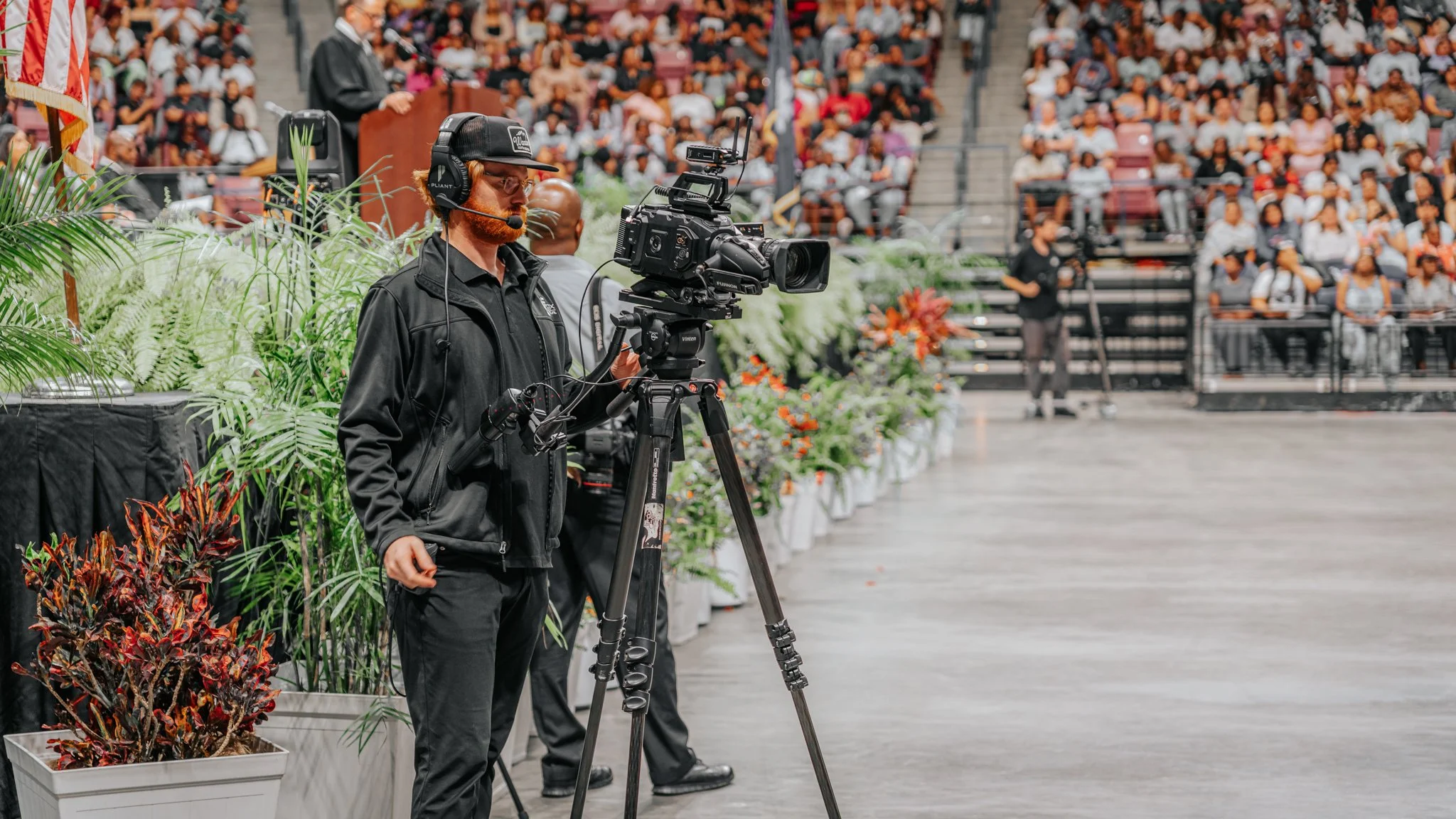 A cameraman using a professional video camera on a tripod at an indoor event with a large seated audience in the background.