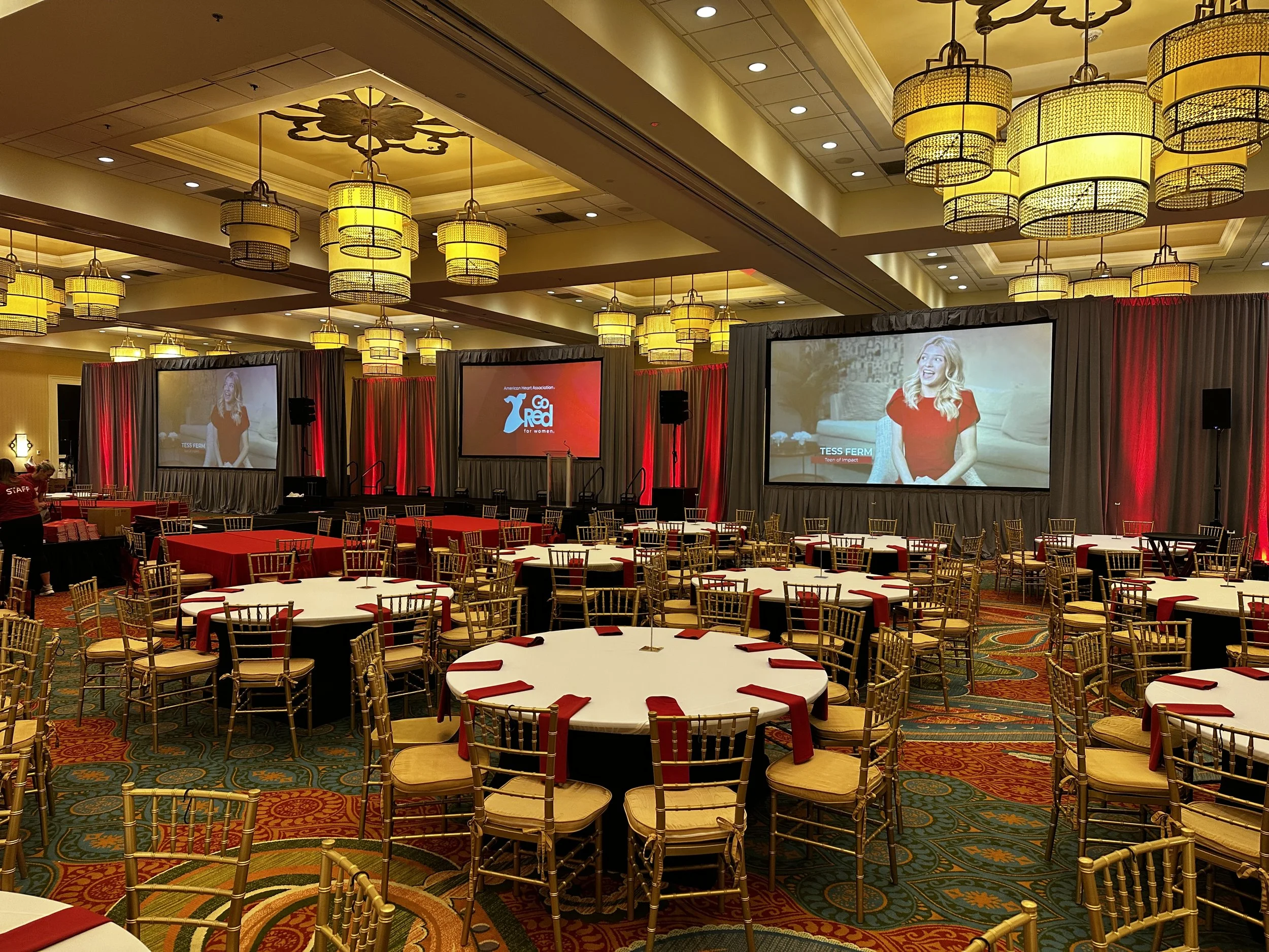 A large banquet hall set up with round tables covered in white tablecloths and decorated with red napkins, surrounded by gold-colored chairs. The hall has high ceilings with multiple large yellow and black hanging light fixtures. In the background, t