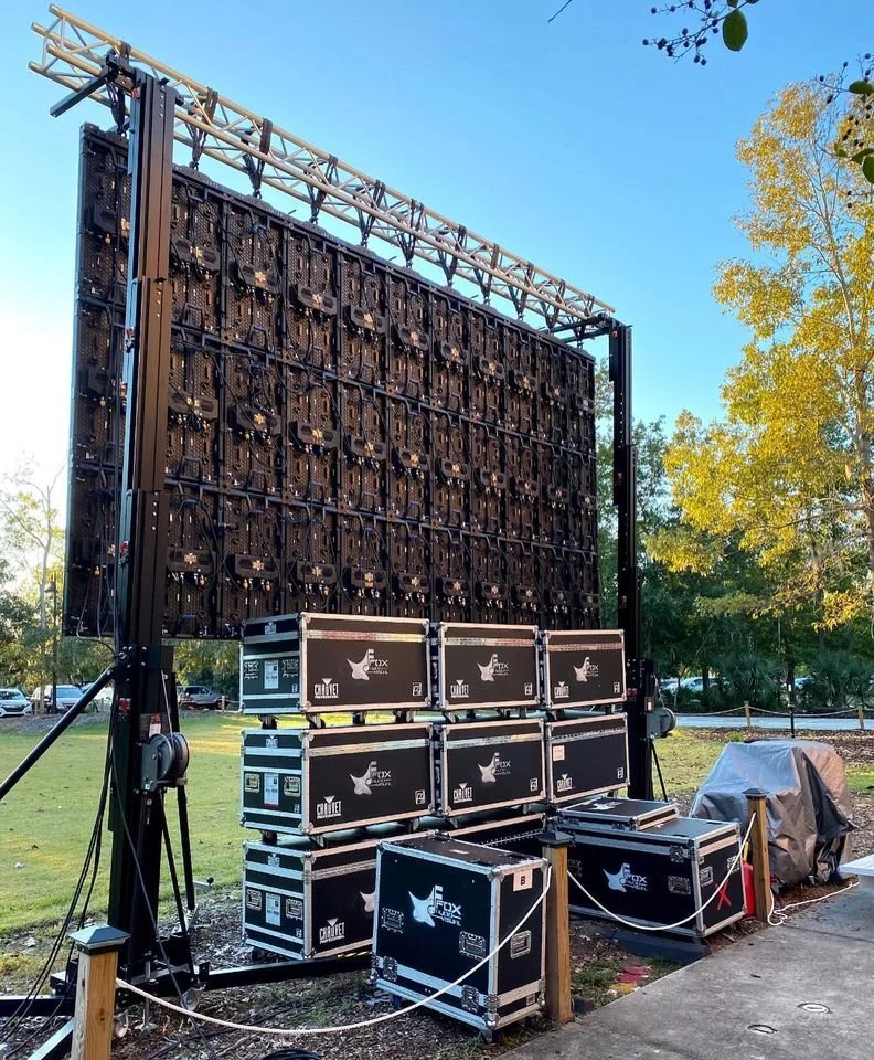Stage lighting and equipment setup outdoors with a large LED screen, black flight cases, and a covered object, surrounded by a wooden barrier and trees.