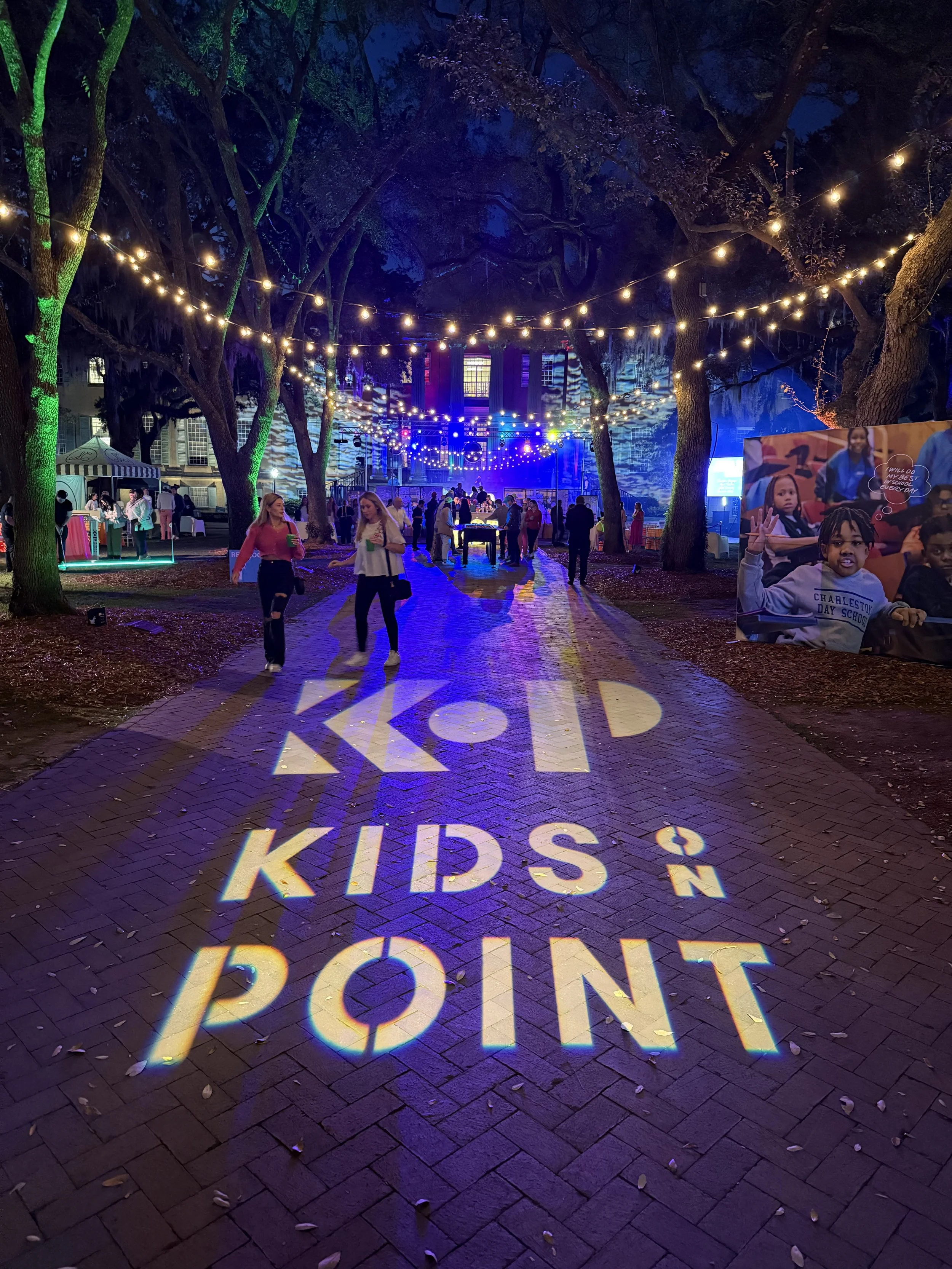 Night scene at a kids' event with string lights hanging overhead, people walking on a brick pathway, and a large sign on the ground that says 'KIDS ON POINT' projected with colorful lights. There are trees surrounding the area and a poster with child