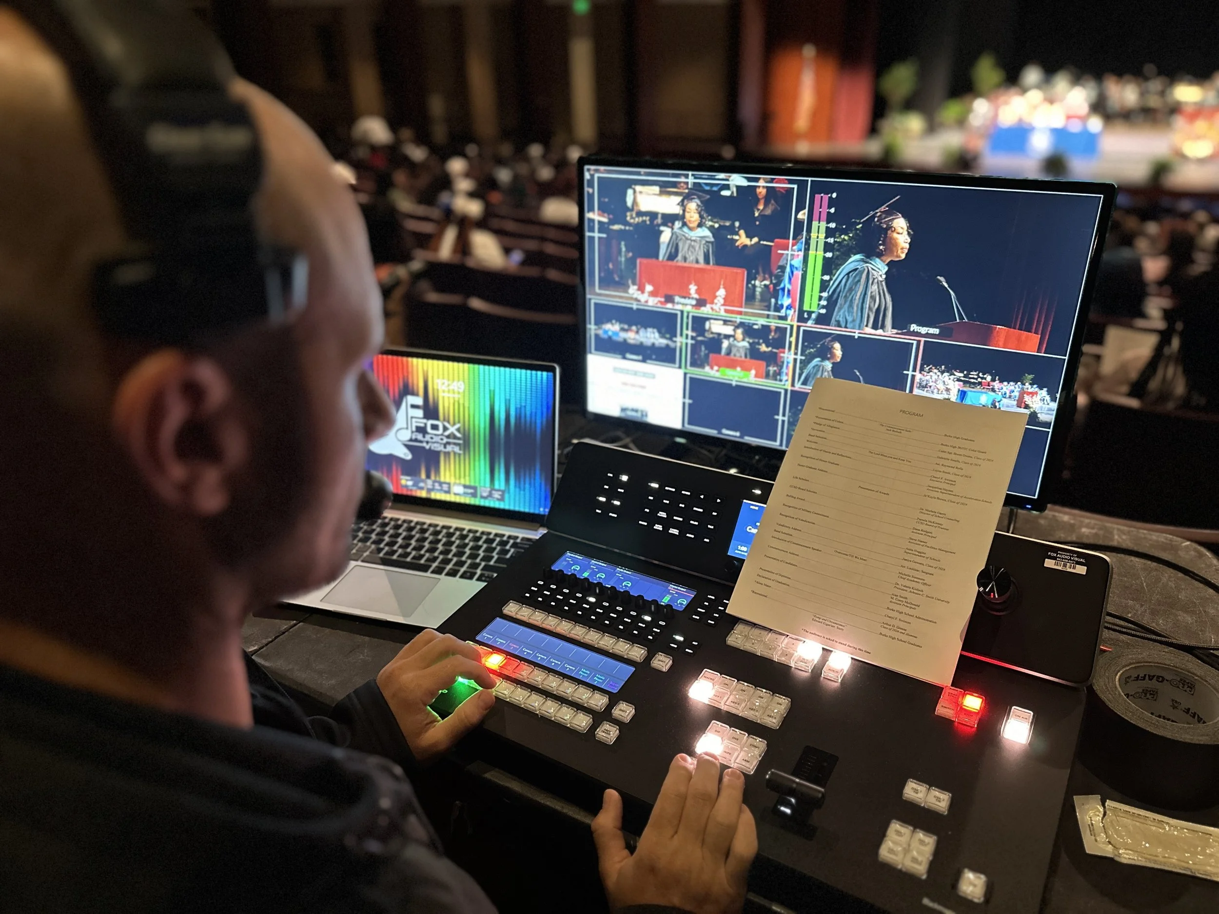 A person operating video and lighting control equipment at an event, with audience members sitting and listening on stage in the background.