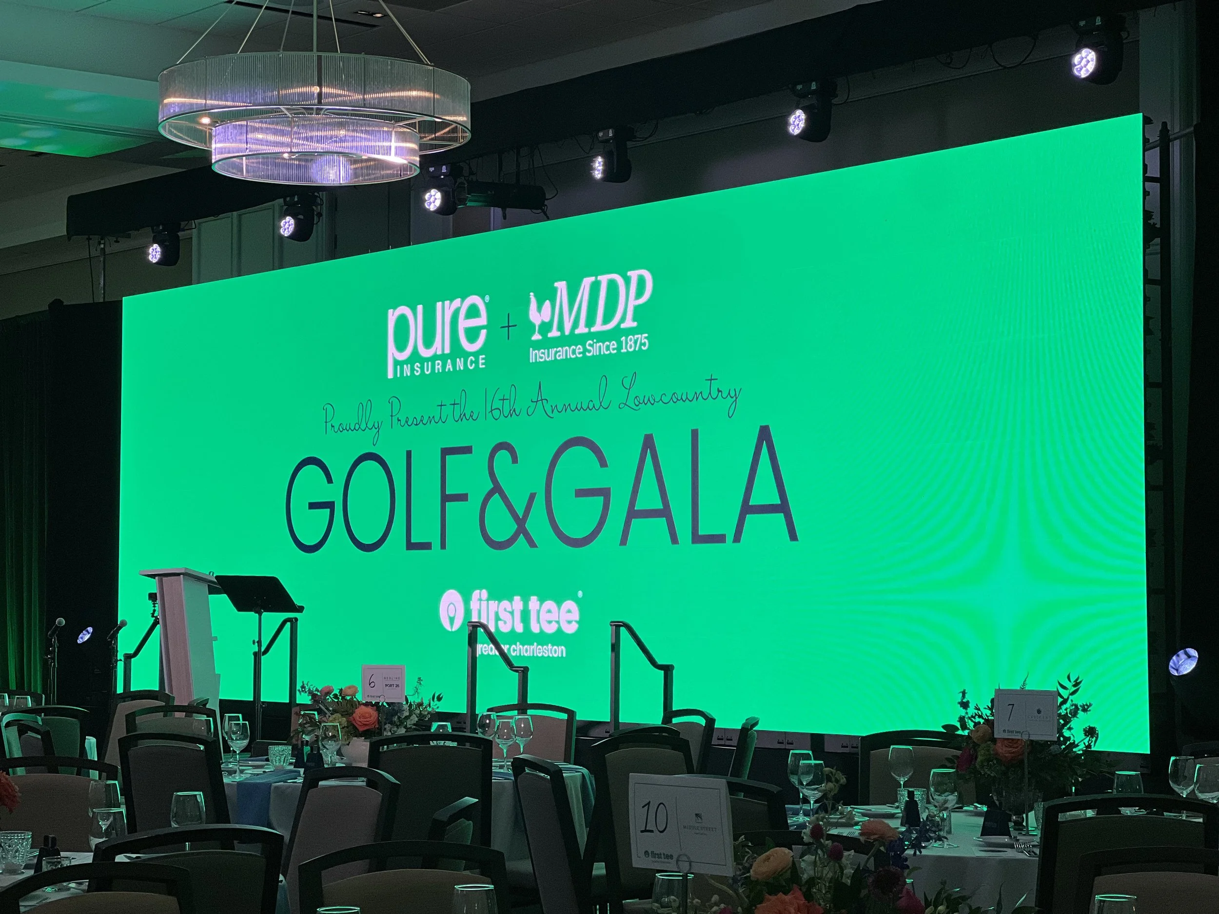 A large green screen with the logos of Pure Insurance and MDVIP, and the text 'Proudly Present the 16th Annual Lowcountry GOLF & GALA' at an indoor event space. There are tables set with flowers, glassware, and place cards, and stage lighting overhea