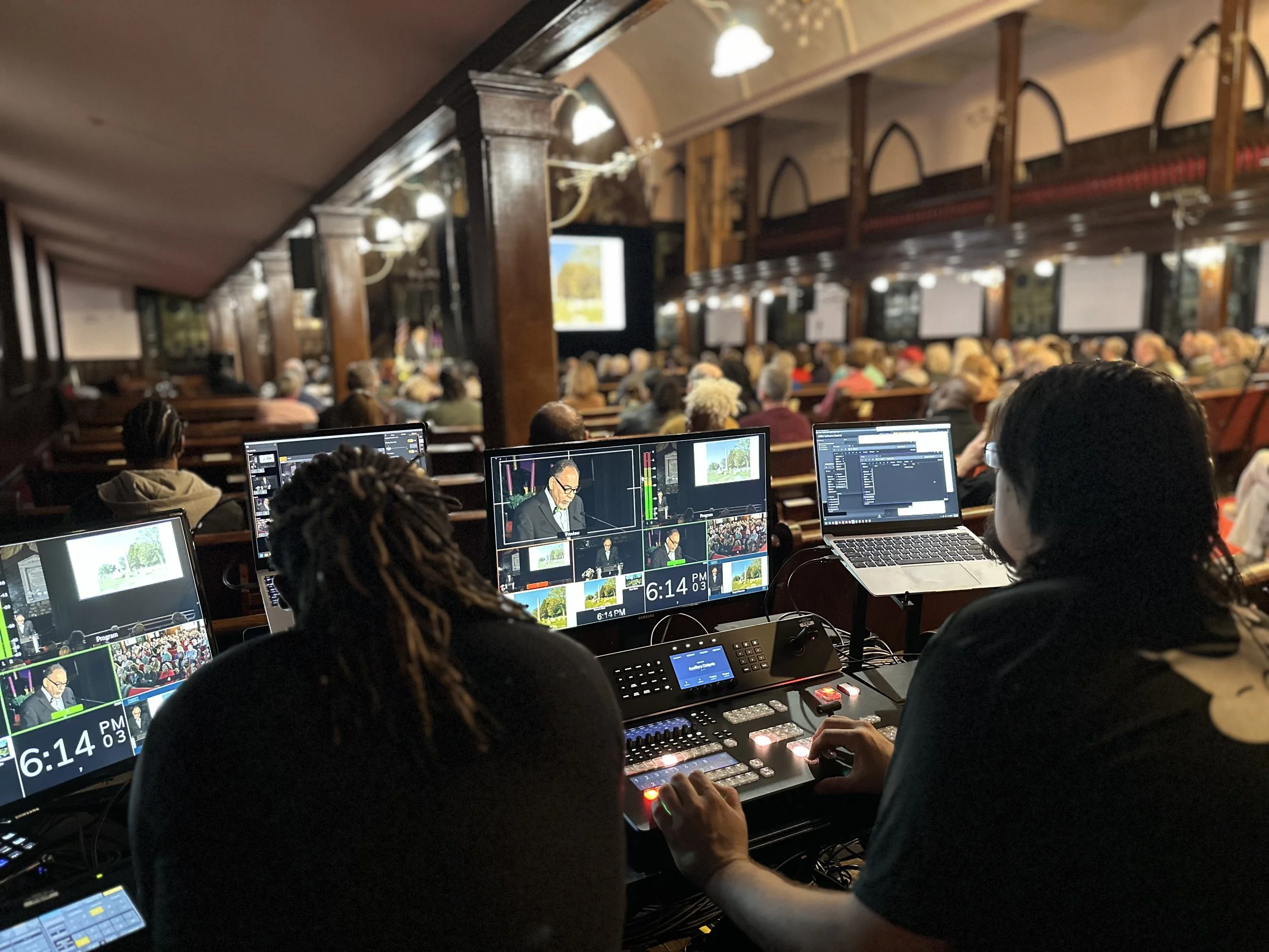 A live event or conference taking place in a large hall with multiple screens, a speaker at a podium, and an audience. In the foreground, there are people operating video and audio equipment with monitors showing the speaker.
