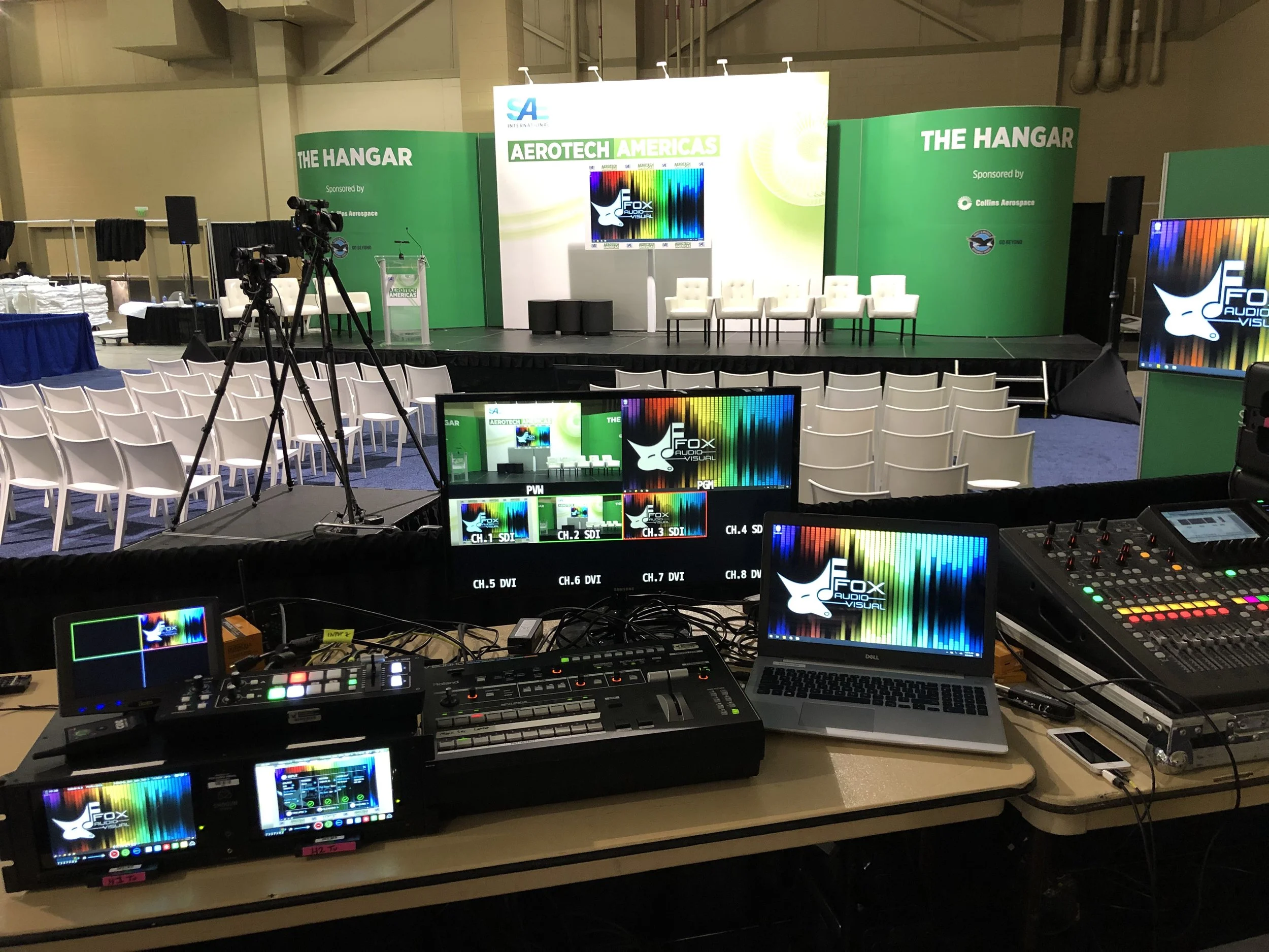 The image shows a stage set up for a conference or event, with a large backdrop featuring green and white colors, and the words 'AEROTECH AMERICAS' and 'THE HANGAR'. There are several chairs on stage, a podium with a microphone, and multiple screens 