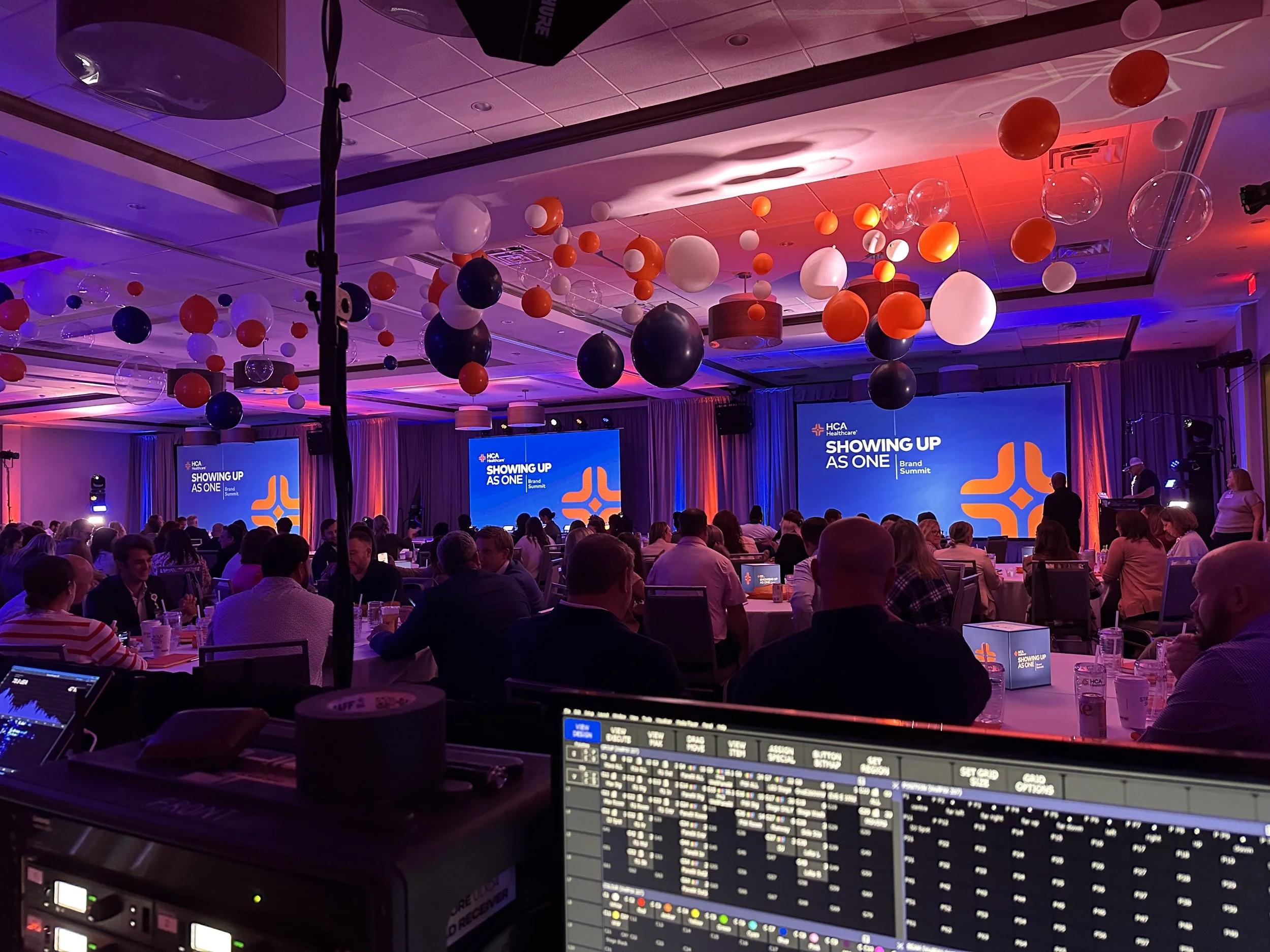 A large conference room filled with seated attendees, a stage with screens displaying the text 'Showing Up As One Brand Summit,' colorful hanging decorations, and a mood lighting setup.